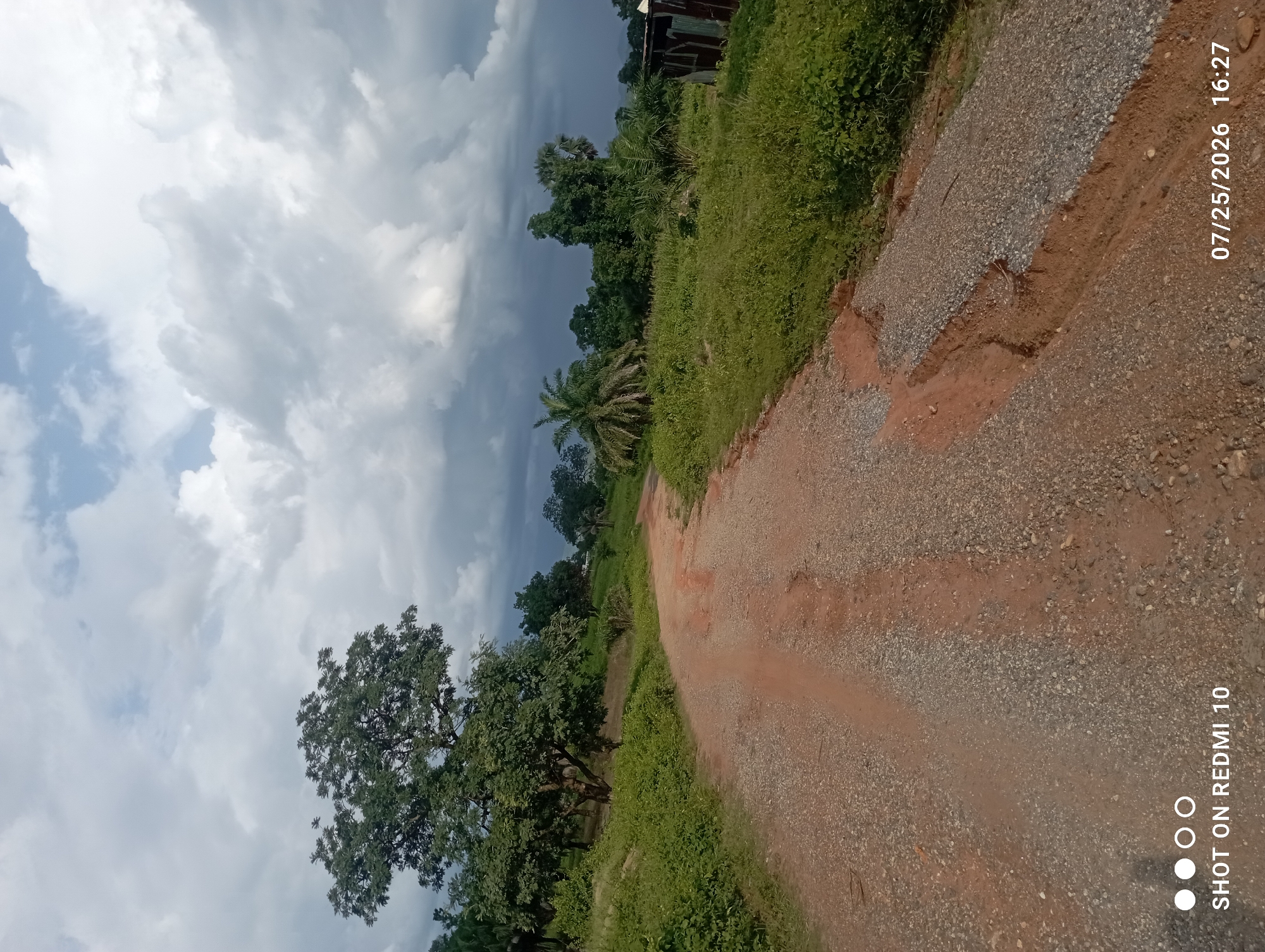 A dirt road curves through a verdant, hilly landscape under a dramatic sky filled with dark, billowing clouds. Lush green vegetation lines the sides of the road, with various trees dotting the scene, including a prominent large tree with dense foliage on the left. In the distance, more trees and a small, dilapidated metal structure are visible. The road itself is unpaved, with a reddish-brown dirt base and gravel scattered across its surface. Erosion is evident in some sections, with exposed earth creating gullies. The overall impression is one of a rural, possibly remote, location. The timestamp in the lower right corner indicates the photograph was taken on July 25, 2026, at 16:27. The text "SHOT ON REDMI 10" appears in the lower left corner, identifying the device used for capture. While the image depicts a landscape, there are no people, animals, or specific buildings identifiable within the scene that would pinpoint its exact location in Serti, Nigeria, beyond it being a rural area within that region.