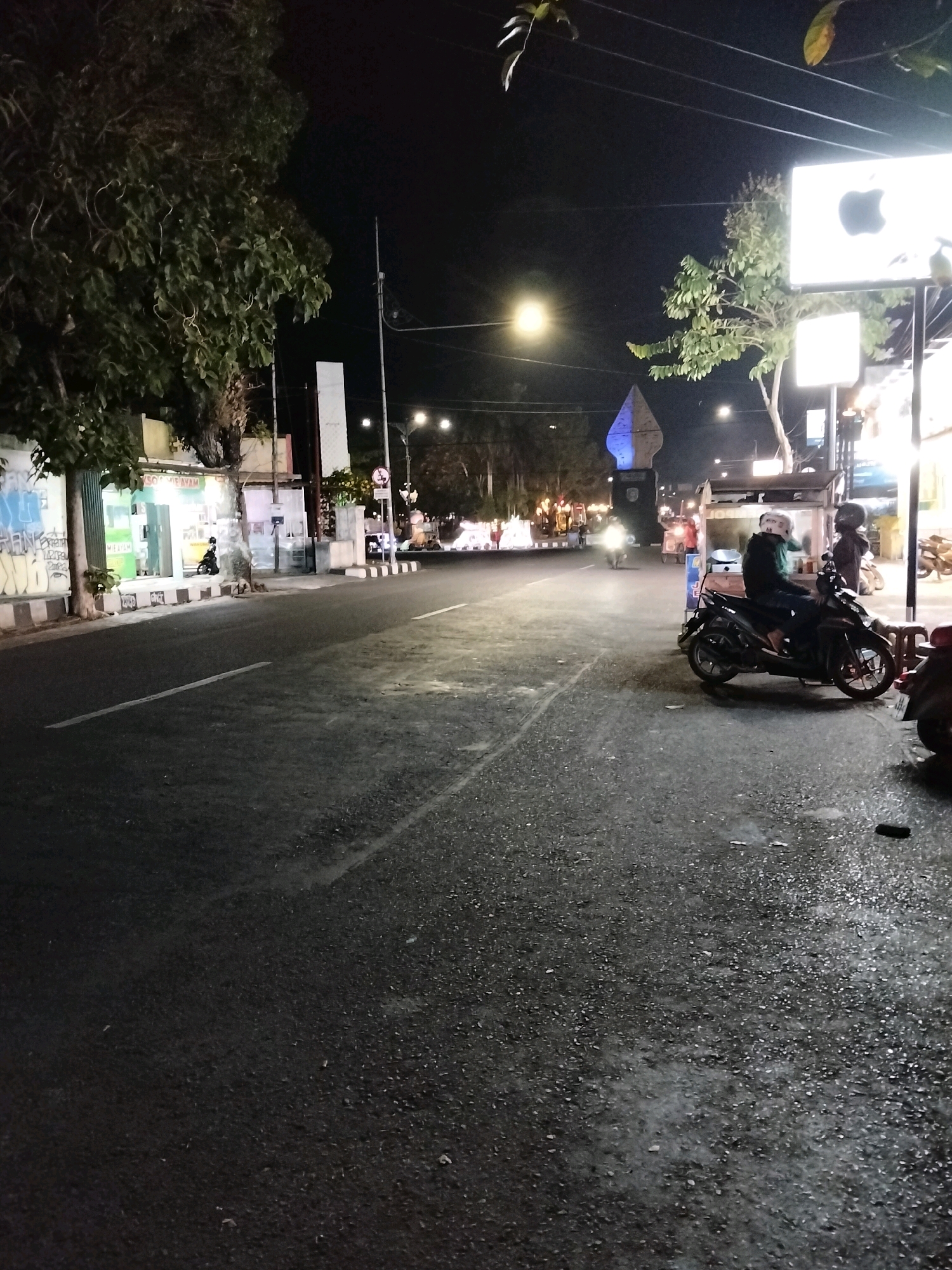 This is a nighttime street-level photograph in the city of Purworejo, Indonesia, likely near a prominent landmark. The scene is illuminated by streetlights and the glowing signs of businesses. On the right side of the road, two individuals are seated on a parked scooter. One person, wearing a green jacket and helmet, is in the driver's seat. Another person, wearing a dark jacket and helmet, is behind them. A food stall is visible behind the scooter.
Further down the street, several other motorcycles and scooters are parked or moving. The road surface appears wet, reflecting the surrounding lights. On the left side of the road, a building with graffiti on its lower section is visible, with trees and utility poles lining the street. In the distance, a tall, blue-lit monument or structure stands out against the night sky. The overall atmosphere suggests a lively commercial or public area at night.