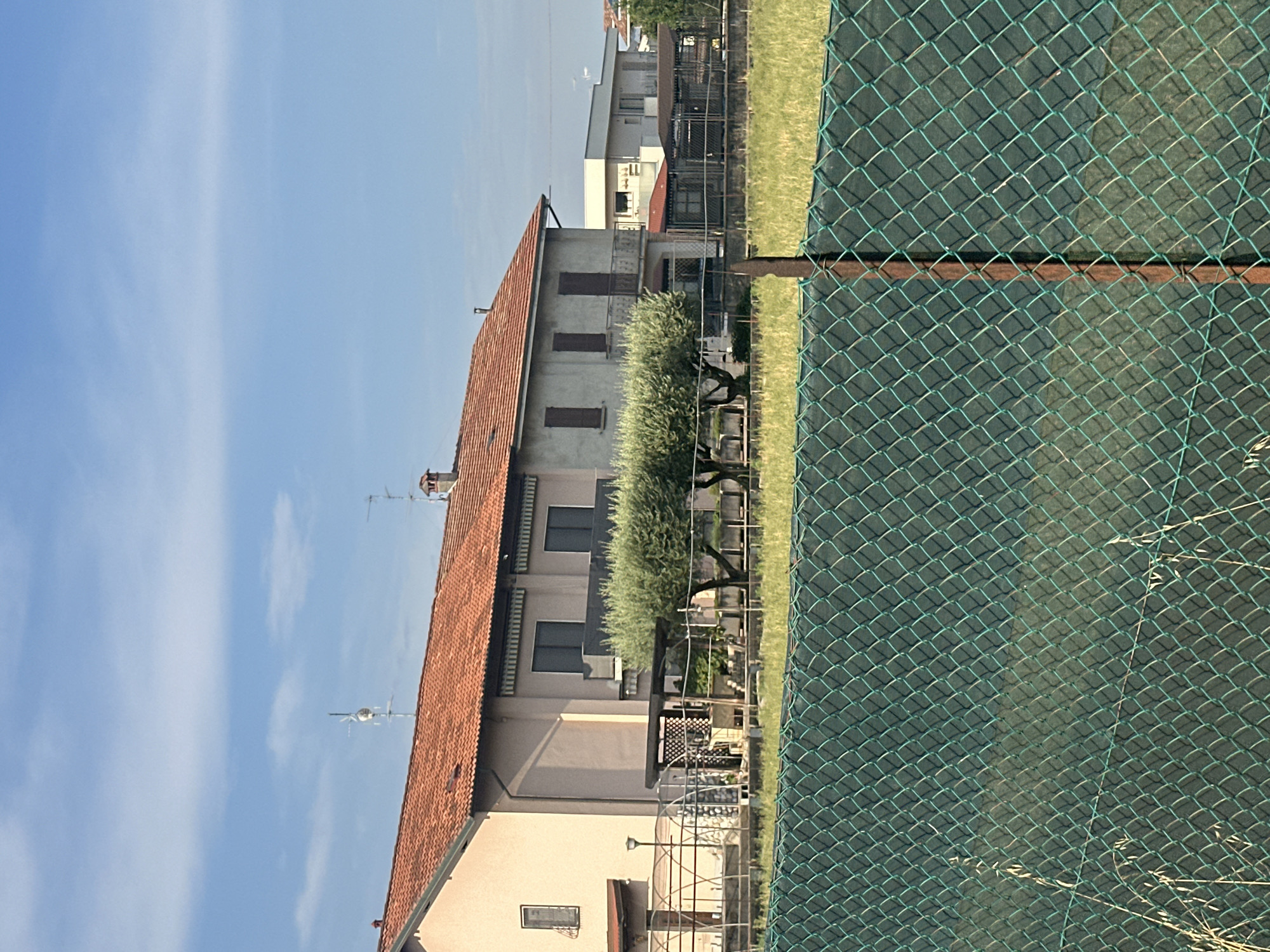 The image depicts a residential area in Chiari, Italy, under a clear blue sky with wispy white clouds. The scene is dominated by a two-story building with a terracotta tiled roof. The building's facade is a light beige color, with dark-framed windows. An olive tree with dense foliage is visible in front of the building. A green chain-link fence runs along the right side of the image, partially obscuring a grassy area and more buildings in the background. The sun appears to be high in the sky, suggesting it is daytime. The overall impression is one of a peaceful, suburban setting. There is no visible text in the image.