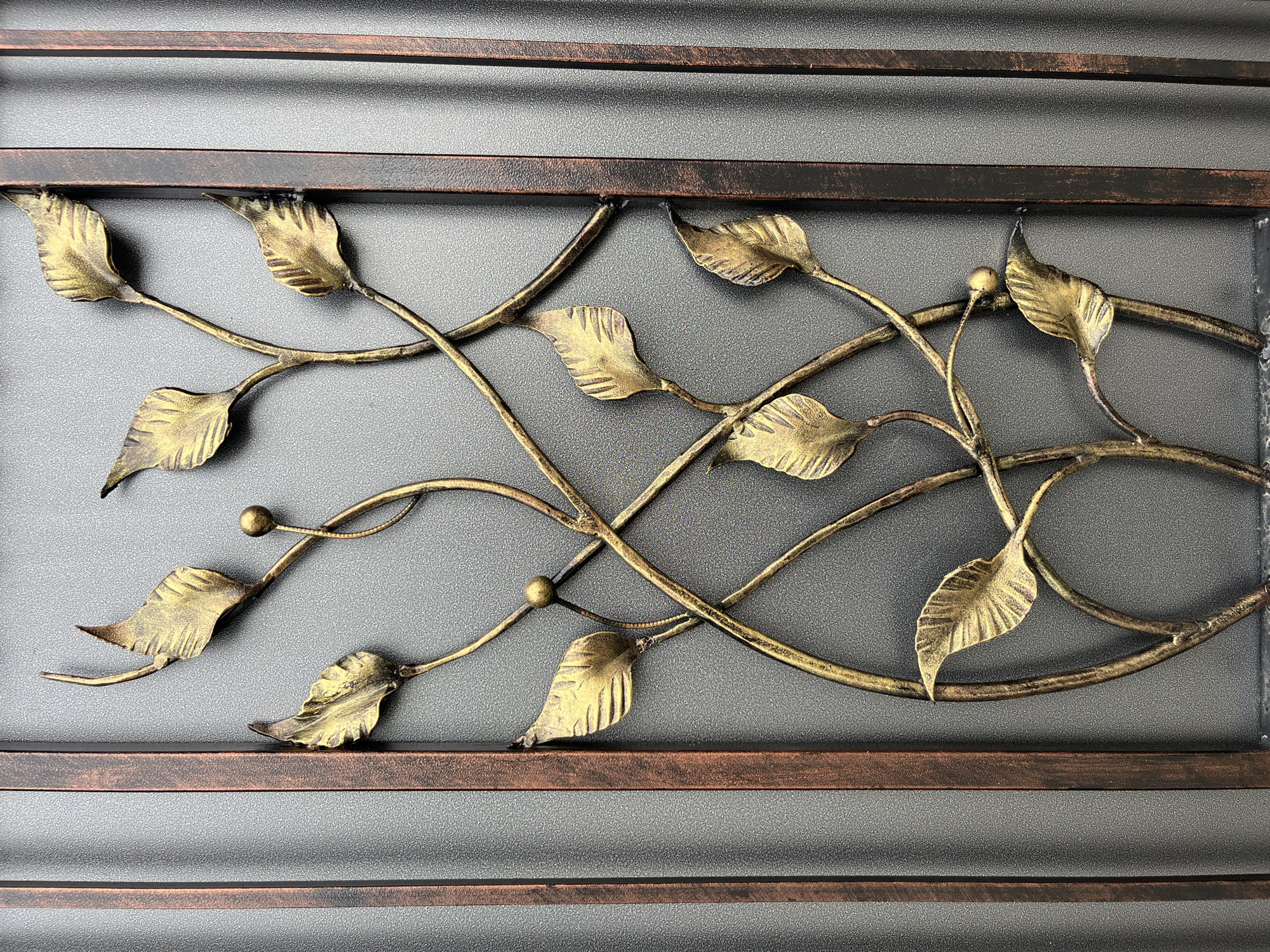 This is a close-up, eye-level, medium shot of a decorative metal gate section. The gate features an ornate design of interwoven bronze-colored branches with golden leaves. The background is a dark gray, textured surface, possibly a wall or another part of the gate. The overall impression is one of intricate craftsmanship. The image does not provide enough information to determine its location in Drohobych, Ukraine.
