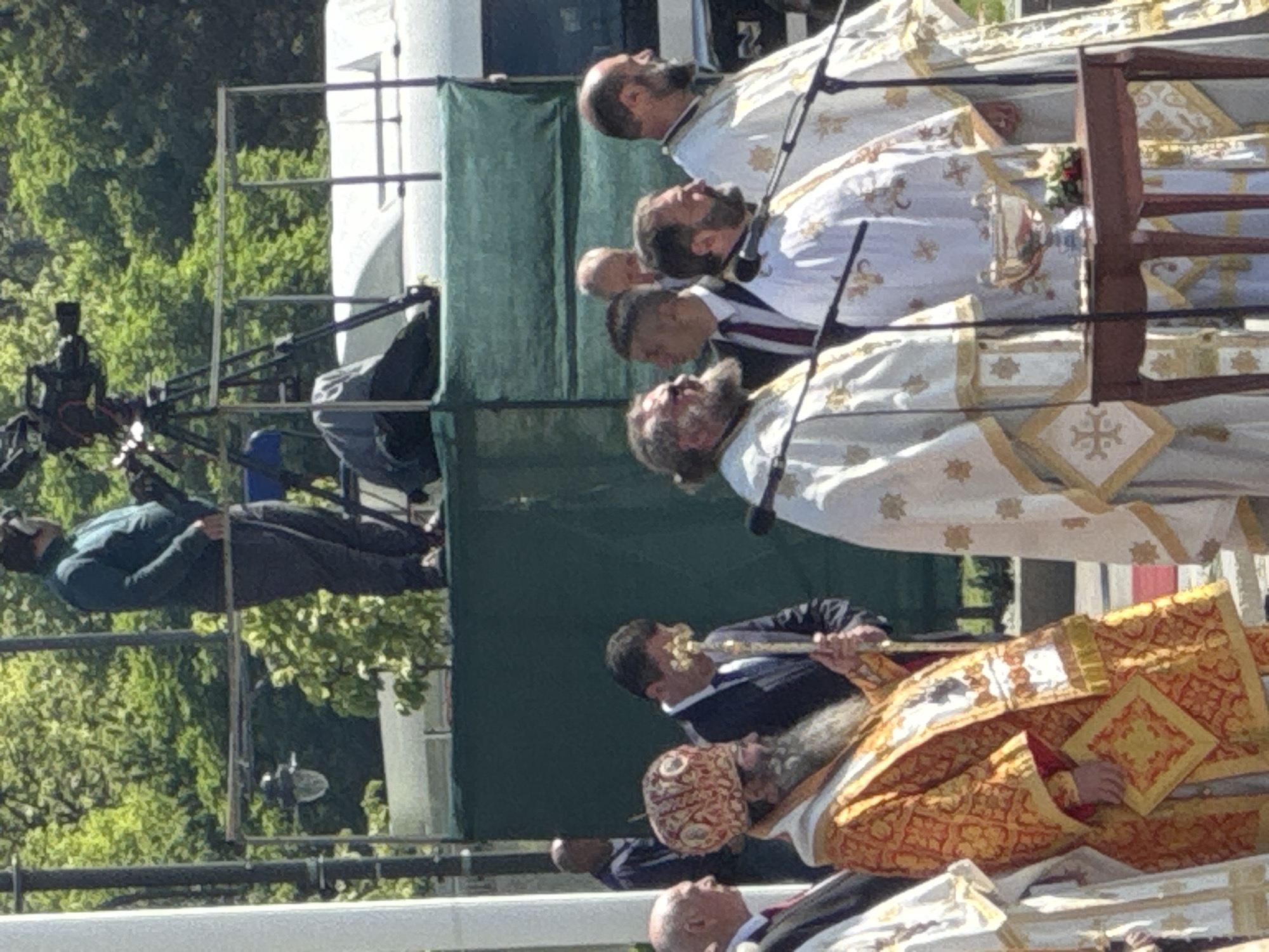 The image shows a procession of Orthodox Christian clergy and laypeople in the city of Sofia, Bulgaria. The clergy are wearing ornate liturgical vestments in white and gold, and one priest is wearing an orange and gold vestment. They are walking with reverence and purpose. Microphones are positioned in front of the group. To the side, a person is seated on a metal structure, with a camera facing the procession. A green screen is in the background, with trees and possibly buildings visible in the distance. A cameraman is visible to the left of the clergy.