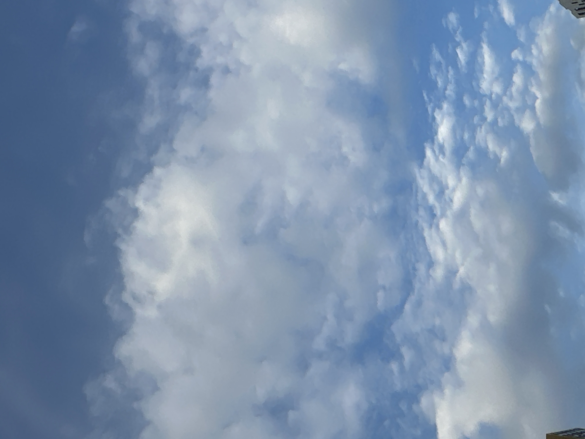 The image shows a blue sky with scattered white clouds. The clouds vary in size and density, with some appearing wispy and others more substantial. The lighting suggests it is daytime, likely late morning or early afternoon, given the brightness of the sky and the illumination of the clouds. There are no people or specific objects clearly visible that would indicate a particular activity or event. The scene is open and natural, focusing solely on the sky. There are no visible text elements within the image. The top right corner of the image shows a small sliver of a building or structure with a patterned surface, possibly indicating an urban or semi-urban setting. However, this element is very small and out of focus. The overall impression is of a clear and somewhat cloudy day.