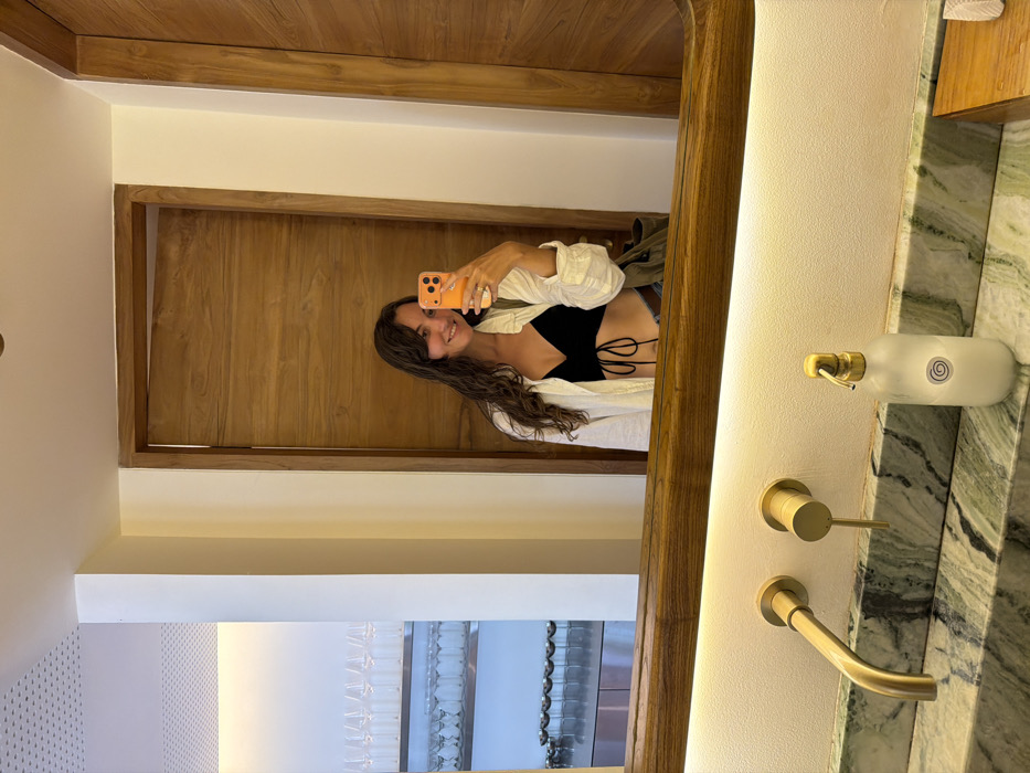 A woman in a black top and white shirt takes a selfie in a bathroom mirror. The mirror reflects a wooden door and frame, a white wall, and a row of glasses on a shelf. The bathroom features a white countertop with green marble accents and gold fixtures, including a faucet and soap dispenser.