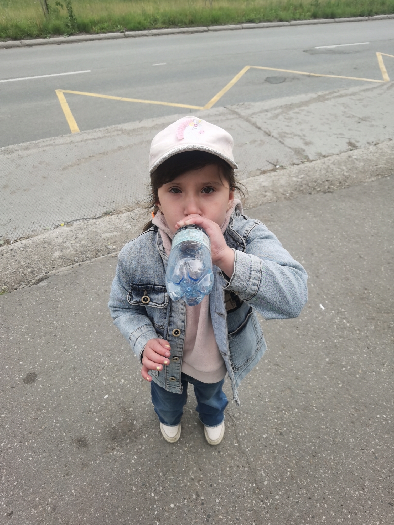 A young girl is depicted outdoors, captured from a high-angle perspective looking down at her. She has dark hair and is wearing a light pink baseball cap with a decorative patch, a light pink hooded sweatshirt, a blue denim jacket, blue jeans, and white sneakers. Her right hand, showing light-colored nail polish with small details, helps hold a clear plastic water bottle to her mouth as she drinks.
The background consists of an asphalt road with prominent yellow and white painted markings. The yellow markings form angular shapes, possibly indicating a no-parking zone or a specific lane designation, while white lines include a solid line and a dashed segment. A concrete curb separates the road from a strip of green grass and sparse vegetation in the distant background. The sky appears overcast, suggesting a cloudy daytime setting.
The girl's expression is neutral, focused on the act of drinking. On the water bottle, "0,3" is visibly printed, likely indicating its volume in liters. The scene conveys a candid moment in an urban or semi-urban environment, possibly along a street or roadside in Chaikovsky, Russia.