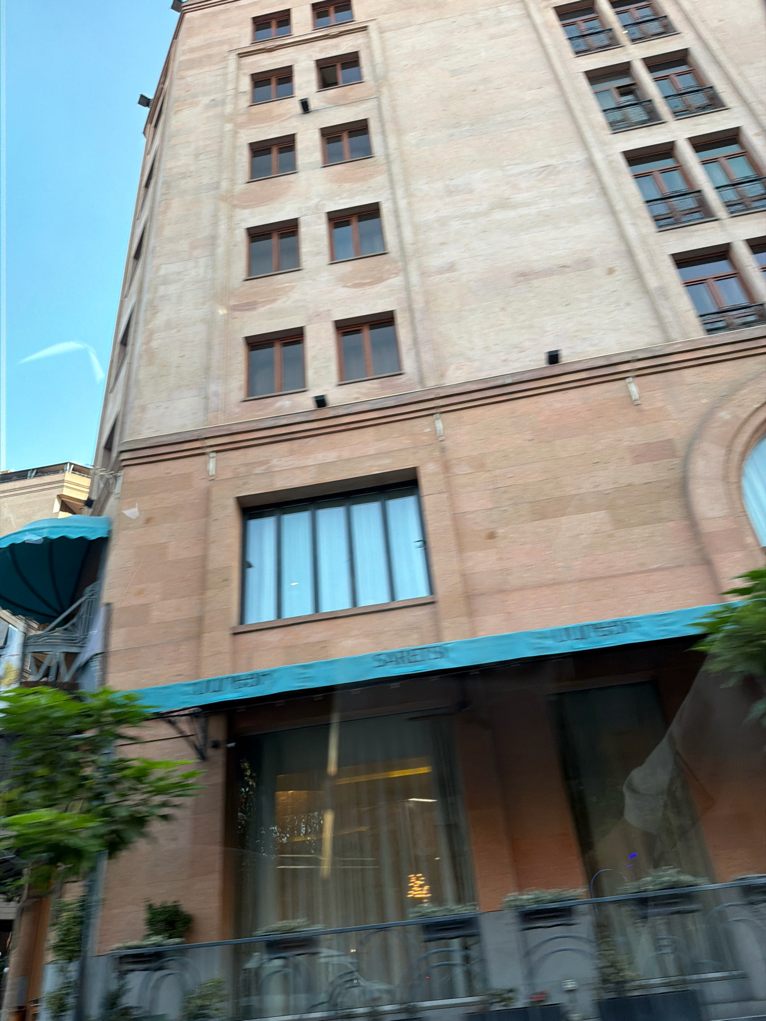 This is a ground-level, slightly upward-angled shot of a multi-story building in Yerevan, Armenia. The building appears to be made of light-colored stone with a darker stone base. The architecture suggests a modern or postmodern style.
The upper portion of the building features numerous regularly spaced, rectangular windows with dark frames. The windows are set into the light-colored stone facade. Some of these windows are open. There are also black square protrusions on the upper facade, possibly lights or ventilation elements.
The lower portion of the building has a different facade material, appearing to be a reddish-brown stone. This section features larger windows with what look like tinted glass, reflecting the sky and possibly interior lighting. A light blue awning extends over a section of this lower facade, supported by metal struts. Text is visible on the awning, though it is partially obscured and blurred by motion.
In the foreground, a green tree with blurred leaves is visible on the left side of the frame, indicating the photo was taken from a moving vehicle or with motion blur. Below the large ground-floor windows, there are decorative planters and metal railings.
The sky is a clear, light blue, with a faint white contrail visible on the left side. The overall impression is that of an urban scene, likely a commercial or residential building in Yerevan.