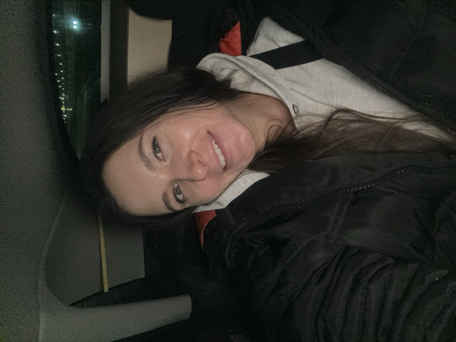 A woman is lying down, likely in a car or confined space, wearing a light gray hooded sweatshirt under a black jacket with an orange accent. She has long brown hair and is smiling. Outside, through a window, lights can be seen at night. The composition is a close-up shot, focusing on her face and upper body, with the surrounding environment appearing dark.