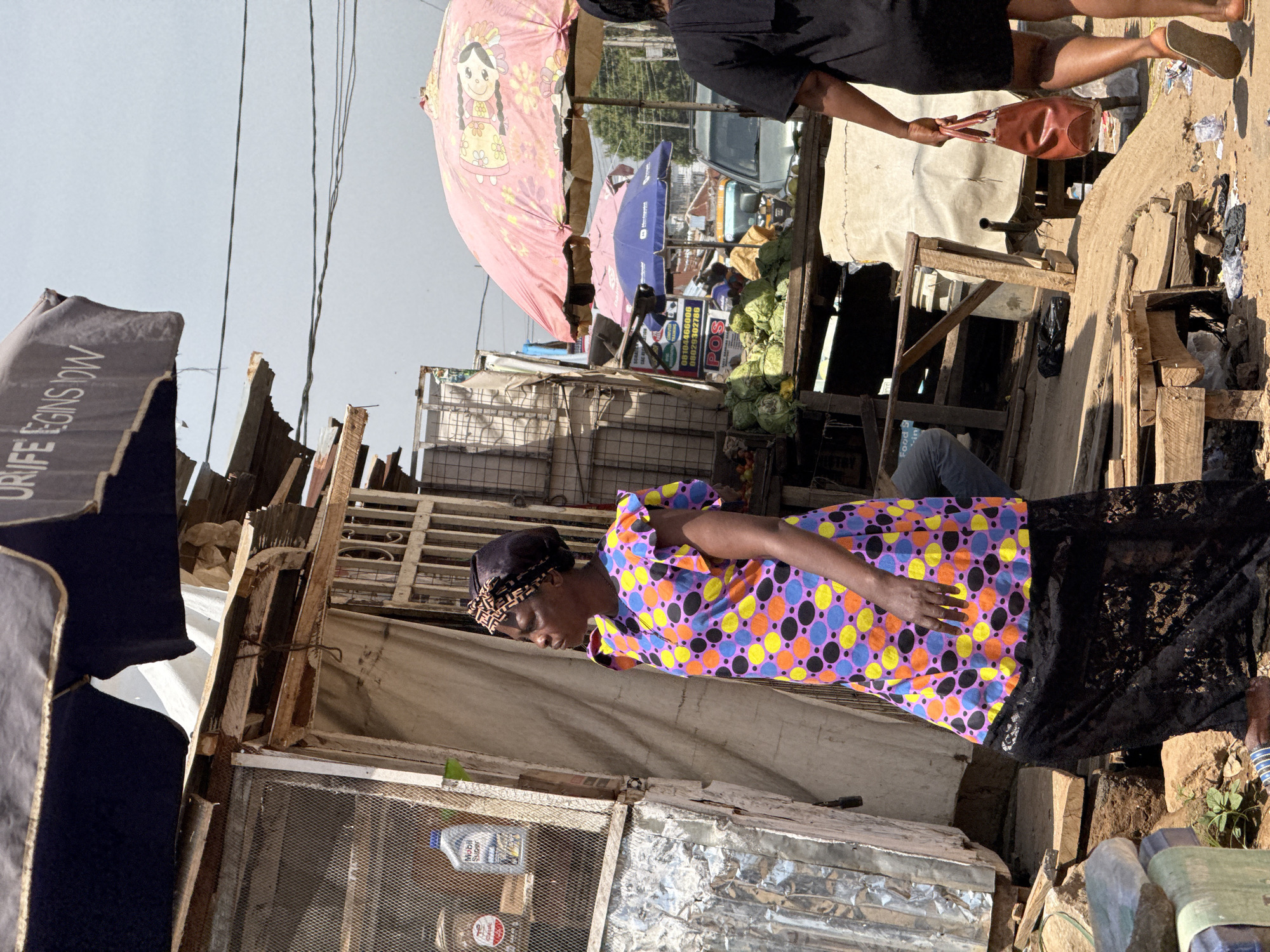 This image depicts a street scene in Abuja, Nigeria, likely a market. A woman wearing a vibrant, multi-colored polka-dot dress and a headscarf stands and looks down, her right hand resting on her stomach. She is wearing a black lace skirt. In the background, a large pink umbrella decorated with a cartoon character is visible, along with a blue umbrella. Structures made of wood and corrugated metal suggest a temporary or informal marketplace. A car is partially visible in the distance, and piles of green vegetables, possibly cabbage, are displayed. The lighting suggests it is daytime, with shadows indicating the sun is present. The scene conveys a sense of everyday commerce. Visible text includes "POS" on a sign and a label on a bottle that appears to be "Mobil Super". The overall impression is one of a lively, albeit somewhat cluttered, outdoor market environment.