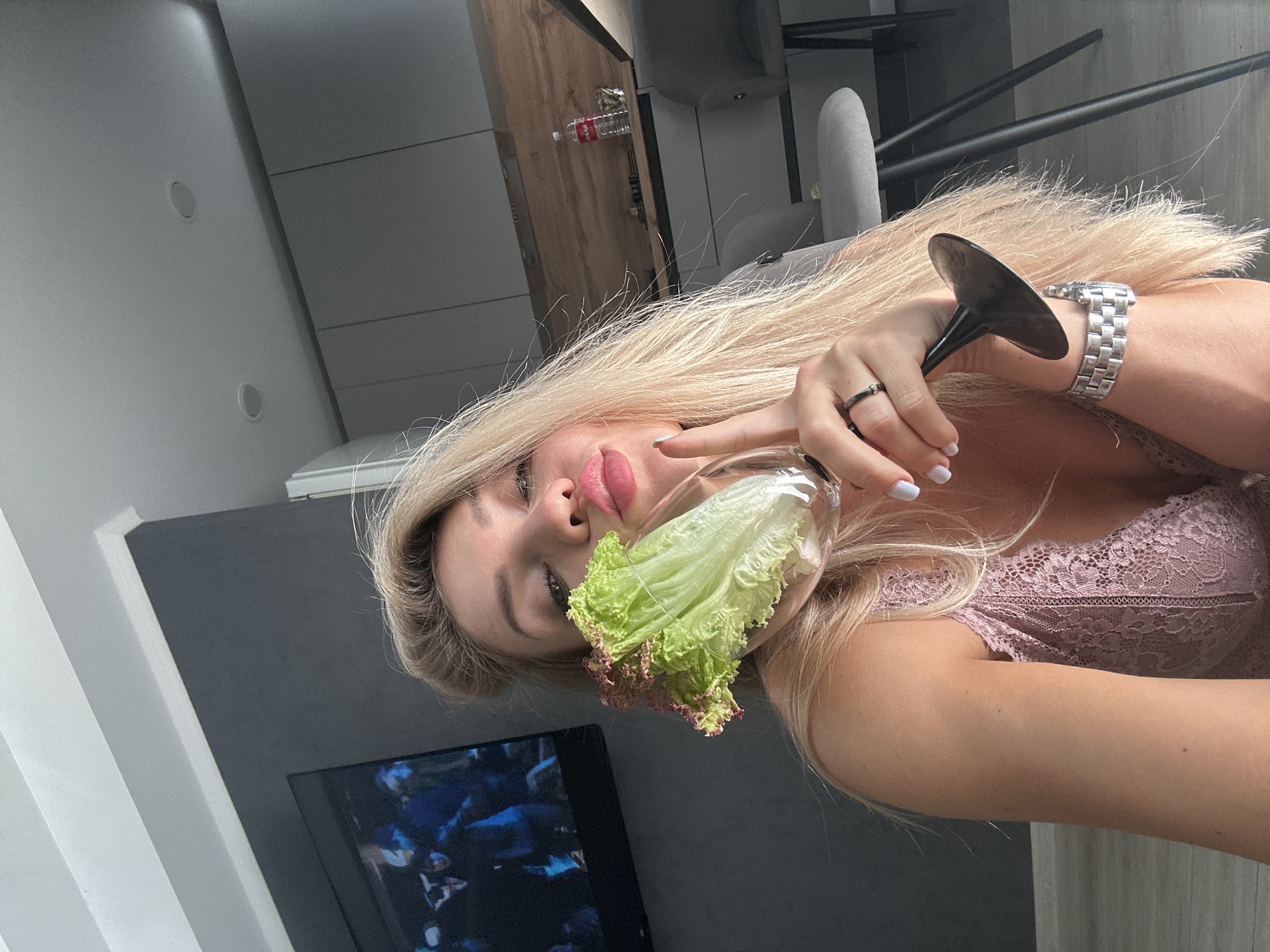 A blonde woman with fair skin and bright pink lipstick holds a wine glass containing a head of lettuce. She is wearing a pink lace bralette and a silver watch. The woman is sitting on the floor in front of a television screen displaying a blue and white image. In the background, a modern kitchen with grey cabinets and a wooden countertop is visible. The scene appears to be indoors.