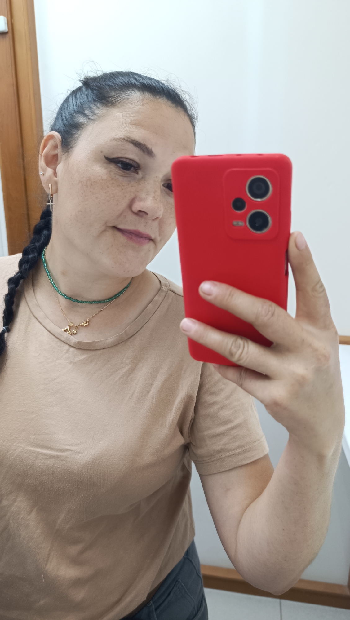 In a medium shot, a woman with dark hair pulled back into a braid, visible freckles, and wearing a tan t-shirt and grey pants, takes a selfie in what appears to be an indoor setting. She is holding up a red smartphone in her right hand, with the camera facing her. She is looking down at the phone screen. Around her neck, she wears two necklaces: a thin green beaded one and a delicate gold chain with a small charm. She has a silver cross earring in her left ear. The background is mostly white, with a wooden doorframe visible on the left. There is no indication of the specific location within the city of Chaikovsky, Russia.