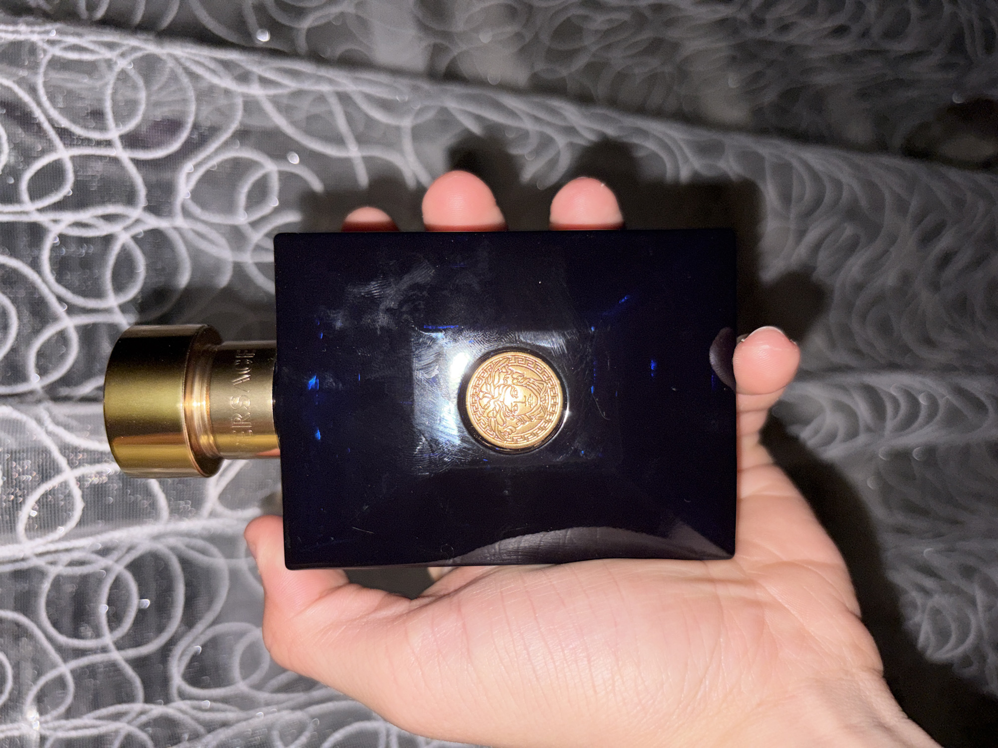 A hand holds a dark blue, rectangular perfume bottle with a gold cap. The bottle has a gold medallion featuring a face, likely a Medusa head, on its front. The background appears to be a draped, textured fabric with a swirling pattern, possibly a curtain. The scene is lit from above, creating highlights on the bottle and the hand. There is no indication of the location being Chistopol, Russia.