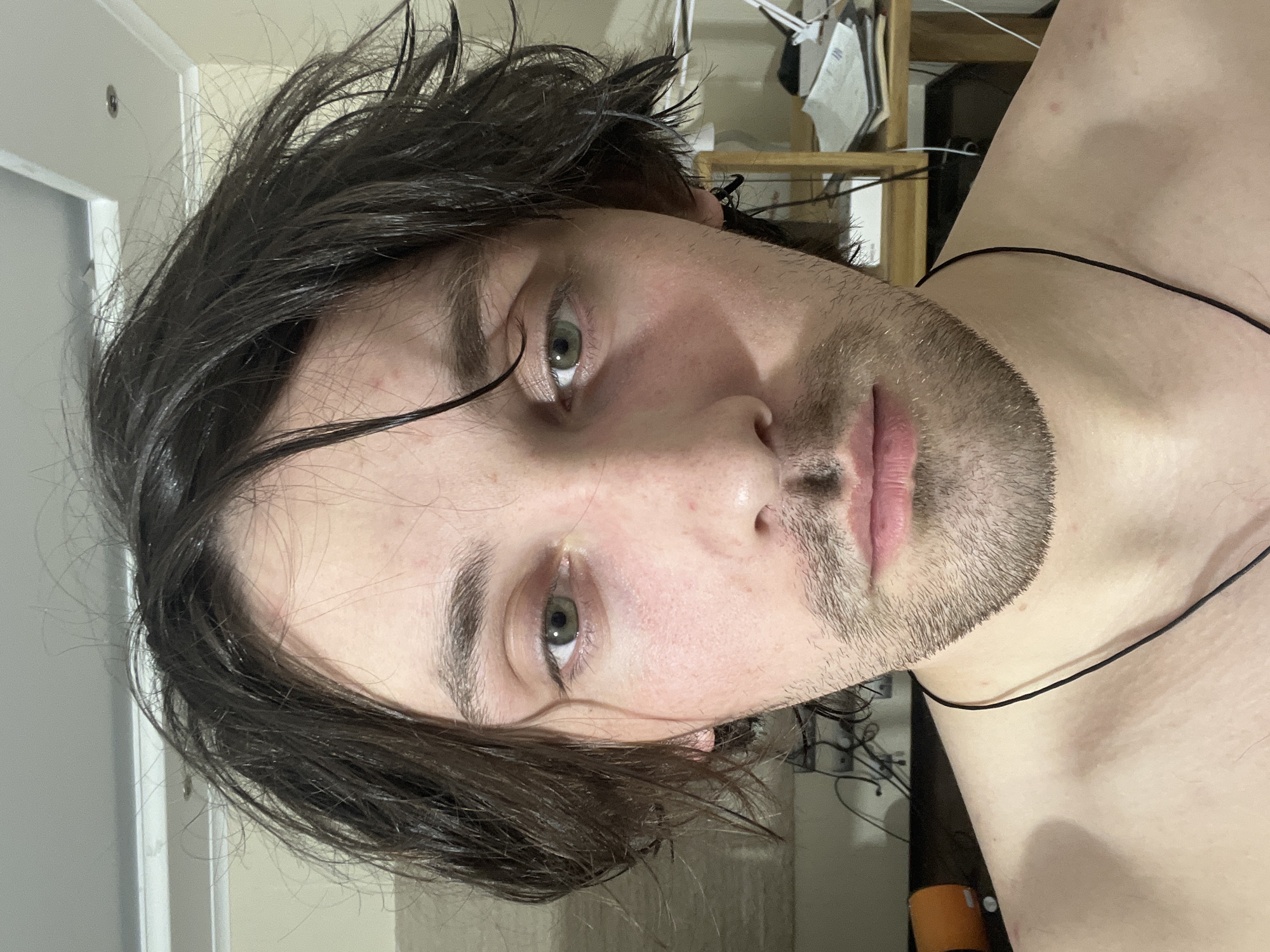 A close-up, slightly rotated, head-and-shoulders portrait of a young man with dark, wet hair and light eyes. He has stubble on his face and a thin, dark cord around his neck. The background is a blurry interior of a room with a white door and beige walls. There's shelving and various items behind him. There is no contextual evidence to suggest the location is Ореховка, Russia.