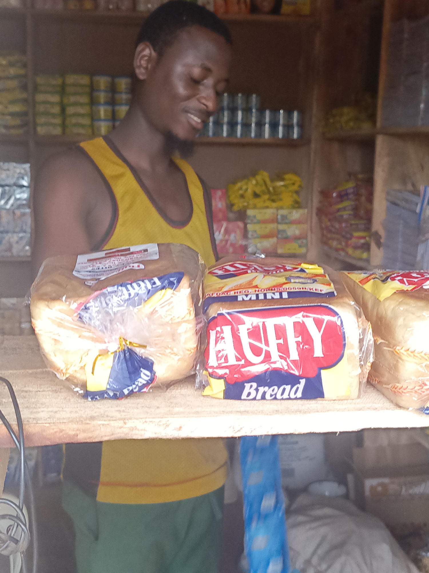 Akwanga, Nigeria: A man in a yellow tank top stands behind a counter in a small shop. On the counter are loaves of bread, with "Huffy Bread" visible on one package. Shelves behind the man are stocked with canned goods and other food items.