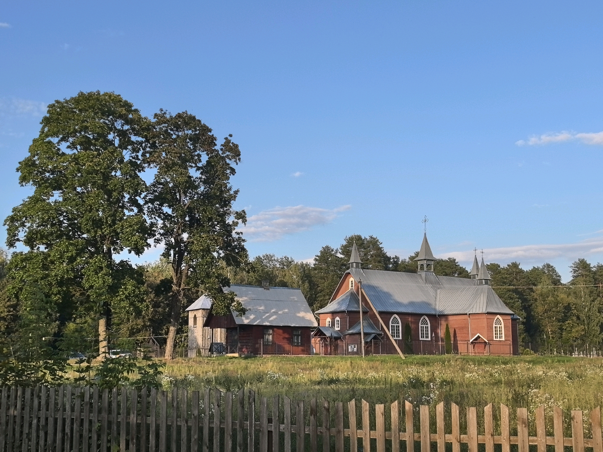 In the city of Парэчча, Belarus, a rustic wooden church with a grey metal roof and a bell tower stands prominently in a grassy field dotted with wildflowers. To the left of the church, a smaller wooden building, possibly a rectory or outbuilding, is attached. Both structures are painted a dark red. A weathered wooden fence runs across the foreground of the image, separating the viewer from the field and buildings. Two large, leafy trees are situated to the left of the church, casting dappled shadows. In the background, a dense line of evergreen trees forms a natural border. The sky is a clear, bright blue with a few wispy white clouds. The scene is illuminated by the warm glow of the late afternoon sun.
