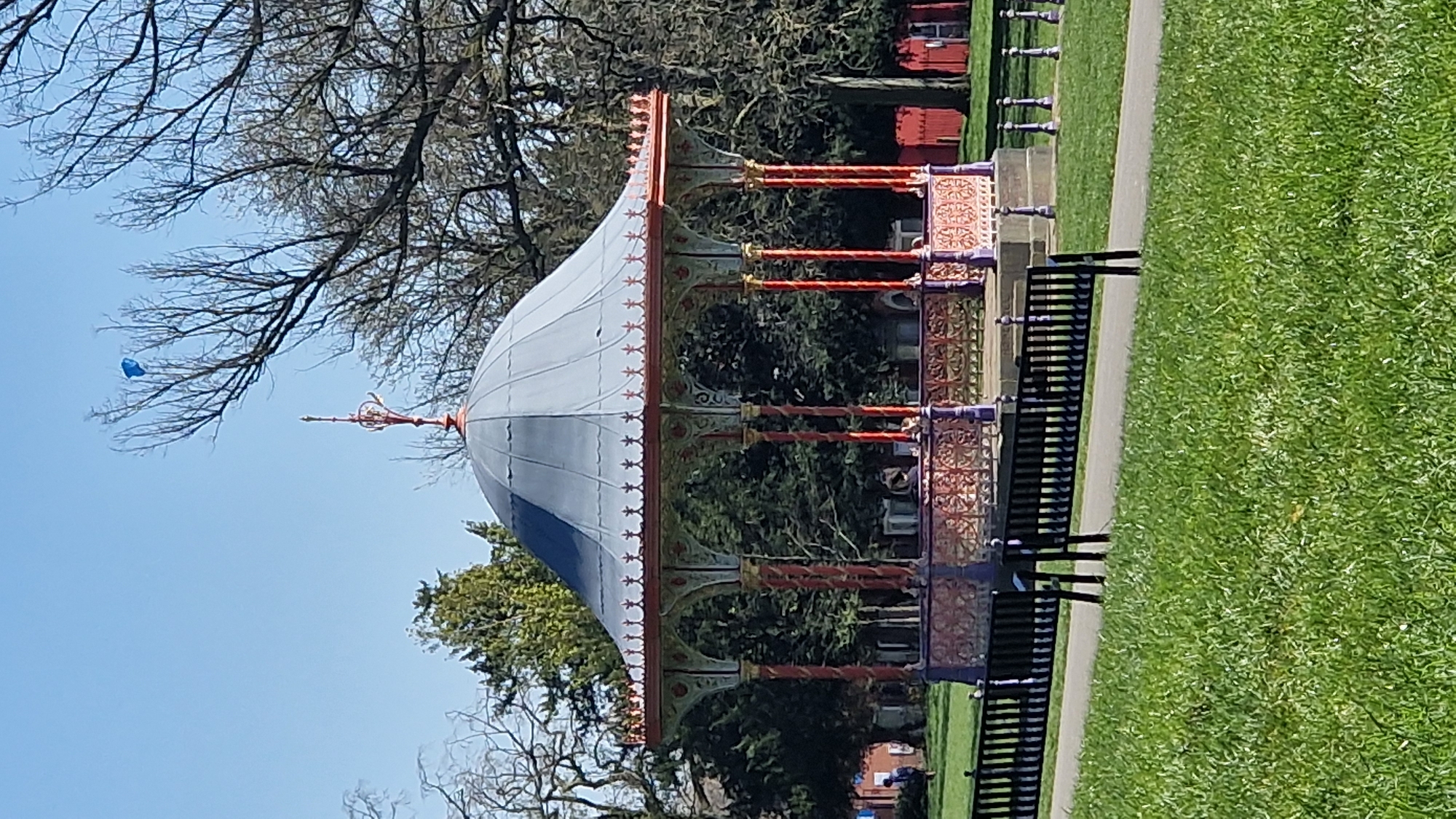 The image shows a Victorian-style bandstand in a park setting. The bandstand has a domed roof, which is white with a blue accent, and ornate metalwork in shades of orange and purple decorates the structure. It is surrounded by green trees and grass. To the right, there is a black bench. The sky is clear and blue. A blue object is visible in the sky, possibly a kite or balloon. The bandstand is located in the city of Lincoln, United Kingdom.