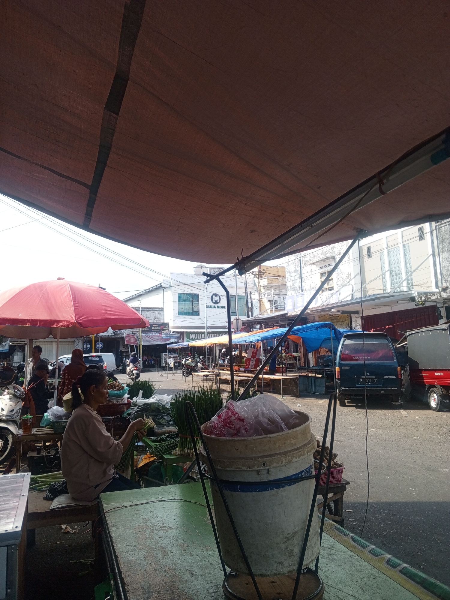 This is an outdoor marketplace scene in Jember, Indonesia, under a large tan tarp. In the foreground, a woman with her hair in a ponytail sits on a wooden bench, engaged in preparing green onions or similar vegetables. She is wearing a light-colored long-sleeved shirt and dark pants. To her right, a large, pale bucket filled with pinkish items wrapped in plastic stands on a metal base. In the middle ground, a red umbrella provides shade, with other vendors and shoppers visible behind it. A white van is parked in the middle of the street. Further back, several stalls are set up with blue and orange tarps. A dark blue minivan is parked on the right side of the street, followed by a red truck. The ground is paved asphalt. The overall atmosphere is bustling and appears to be during daylight hours, with clear skies visible above.