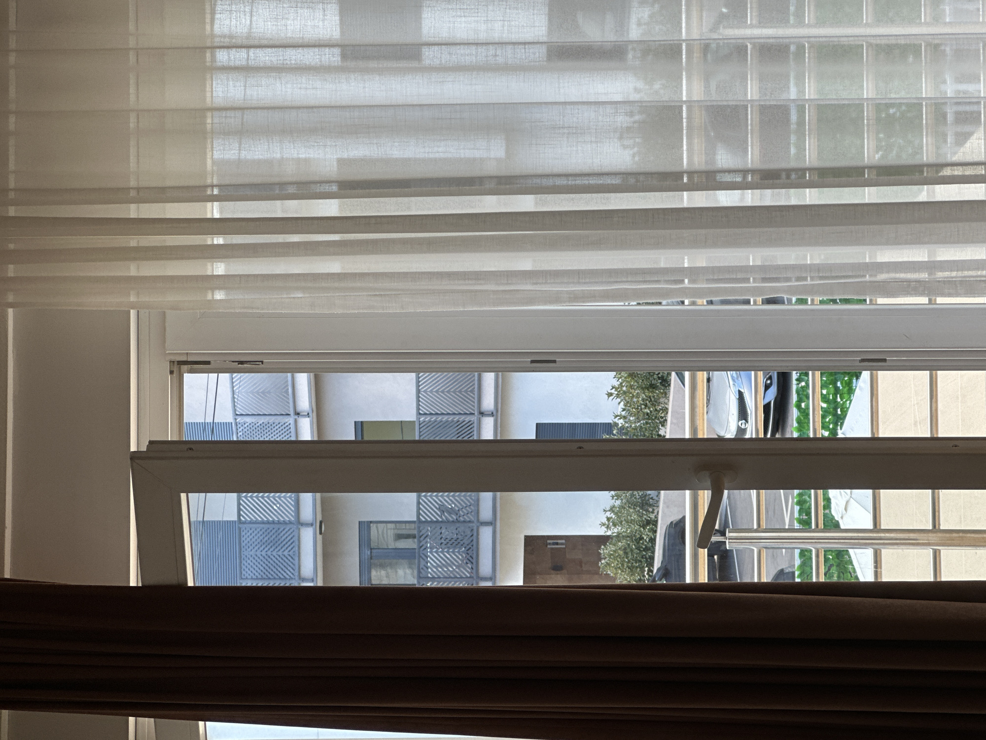 An eye-level, indoor shot looks out an open window with white sheer curtains, and brown pleated curtains at the bottom. Through the open window, a building with diagonally striped window coverings is visible. To the right is a clothesline with laundry hanging on it. The location appears to be in a densely populated urban area. The location of Bar, Montenegro, is not visually identifiable in this image.