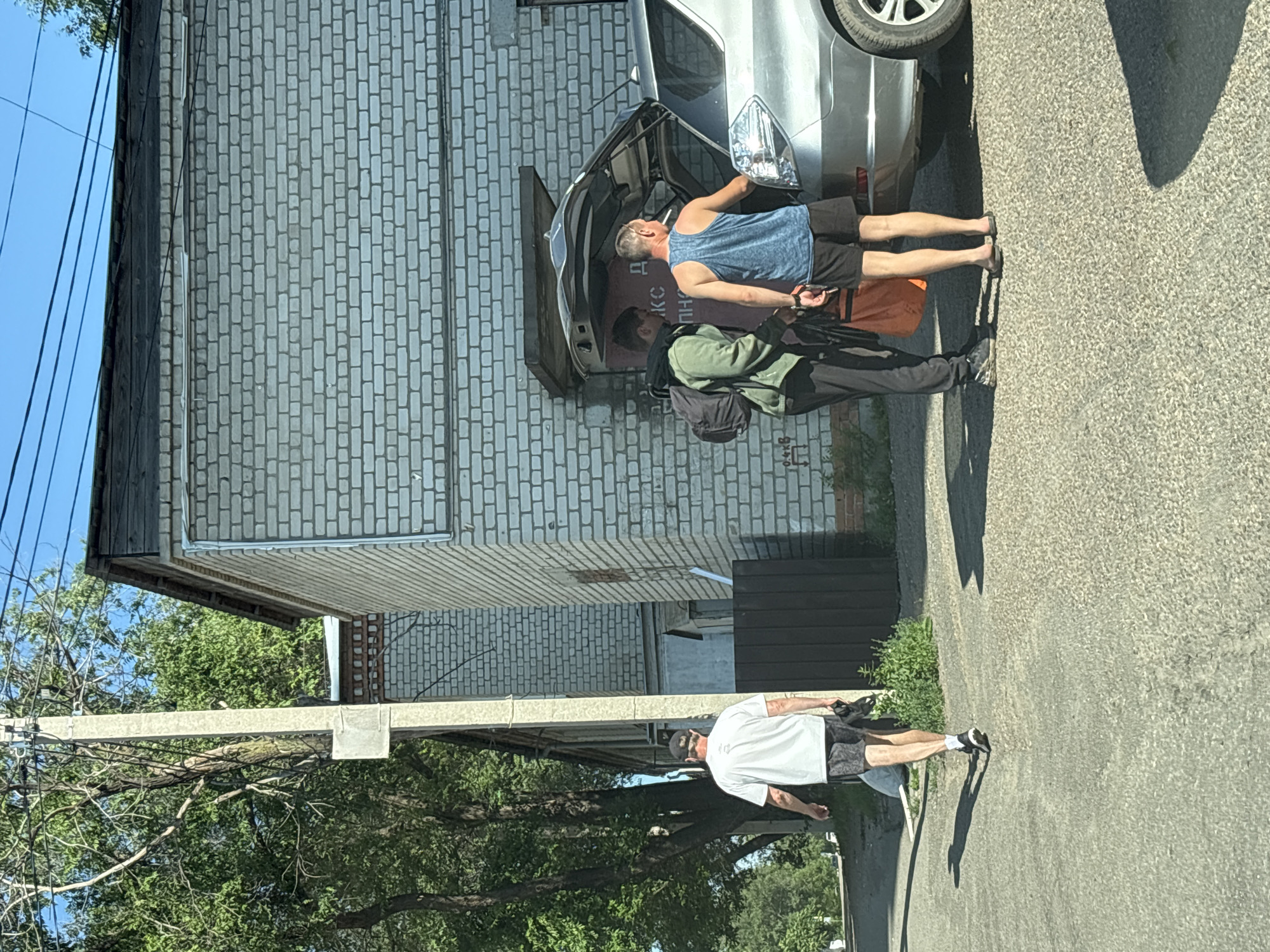 The image depicts an outdoor scene in Blagoveshchensk, Russia, on a sunny day with a clear blue sky. In the foreground, a man wearing a white t-shirt, black shorts, and a black baseball cap is walking away from the camera, holding a black bag in his right hand. His left arm is down by his side. He is on a paved surface that appears to be a road or a parking area.
To the right of the walking man and closer to the camera, two other individuals are standing next to an open car trunk. One is a young man with a backpack, wearing a green hooded jacket and dark pants. He is holding a large orange bag. The second individual is an adult male wearing a blue tank top and black shorts, with his back to the camera. He appears to be reaching into the car trunk. The car is a silver SUV.
Behind the individuals and the car is a light-colored brick building with a dark wooden roofline. To the left of the building and further back is a tall utility pole with wires running across it. Trees with green foliage are visible in the background, casting shadows on the ground. The overall environment suggests a semi-urban or industrial area.