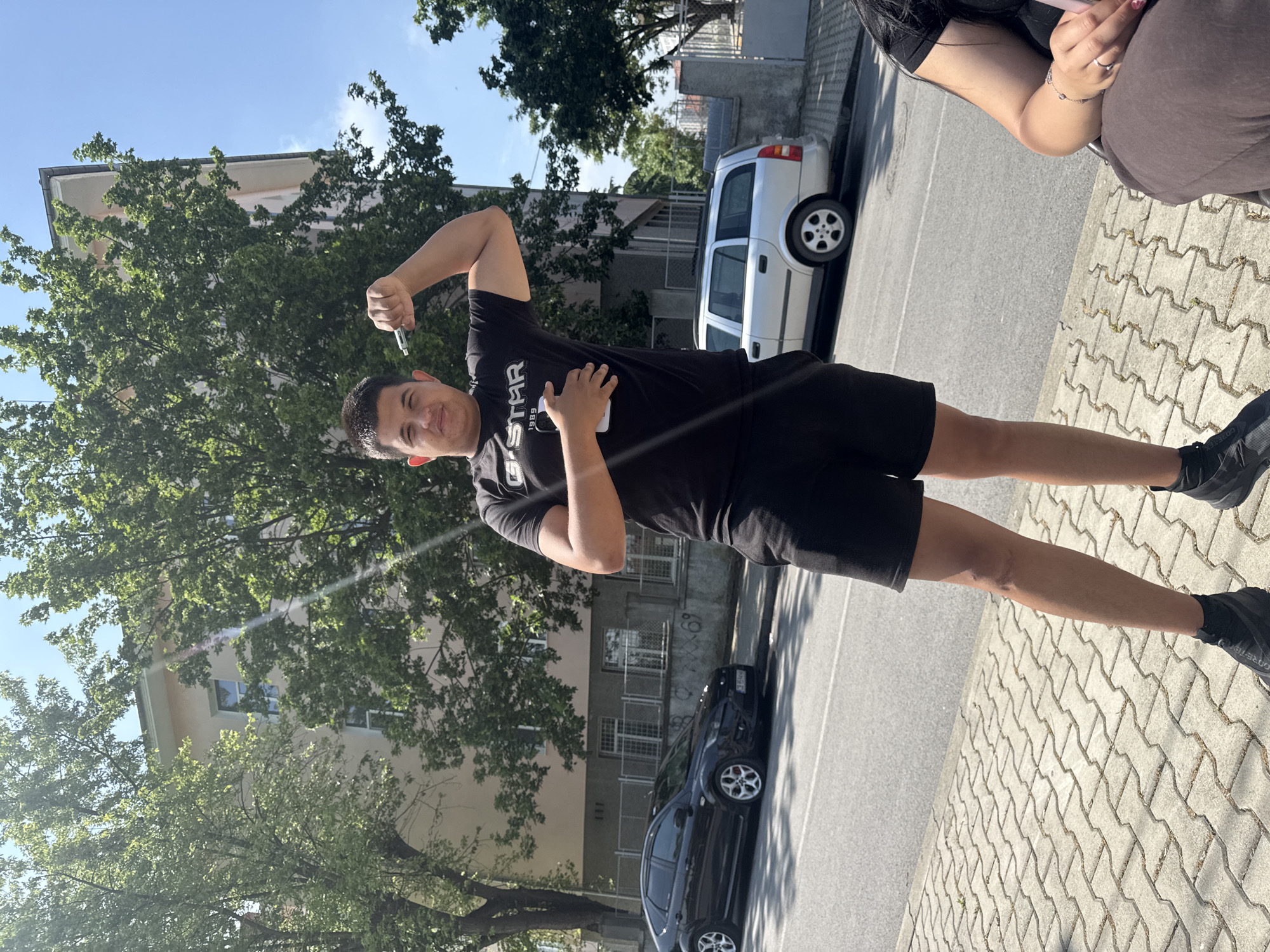 A young man poses for a photo on an urban street during bright daylight. He is standing upright, facing the camera with a playful smile, and has his right arm bent, appearing to flex. In his right hand, he holds a small, pen-like object, possibly a vape pen. His left hand holds a white rectangular object, possibly a smartphone or another vape device, pressed against his chest. He wears a black T-shirt with the text "G-STAR" prominently displayed, and "1989" below it. He also wears black shorts and black athletic shoes, on which "GORE-TEX" is faintly visible.
Partially visible on the right side of the frame is another person, likely female, whose arm, hand, and a portion of her torso are visible. She wears a brown outfit and has rings on her fingers and a bracelet on her wrist.
The setting is an outdoor street scene with a paved sidewalk on the right, featuring interlocking concrete blocks, and an asphalt road on the left. Two cars are parked on the street: a silver station wagon with a roof rack and a black sedan. In the background, residential buildings with windows are visible, interspersed with numerous green trees under a clear, bright blue sky, indicating sunny weather. The scene suggests a casual moment in Elhovo, Bulgaria.