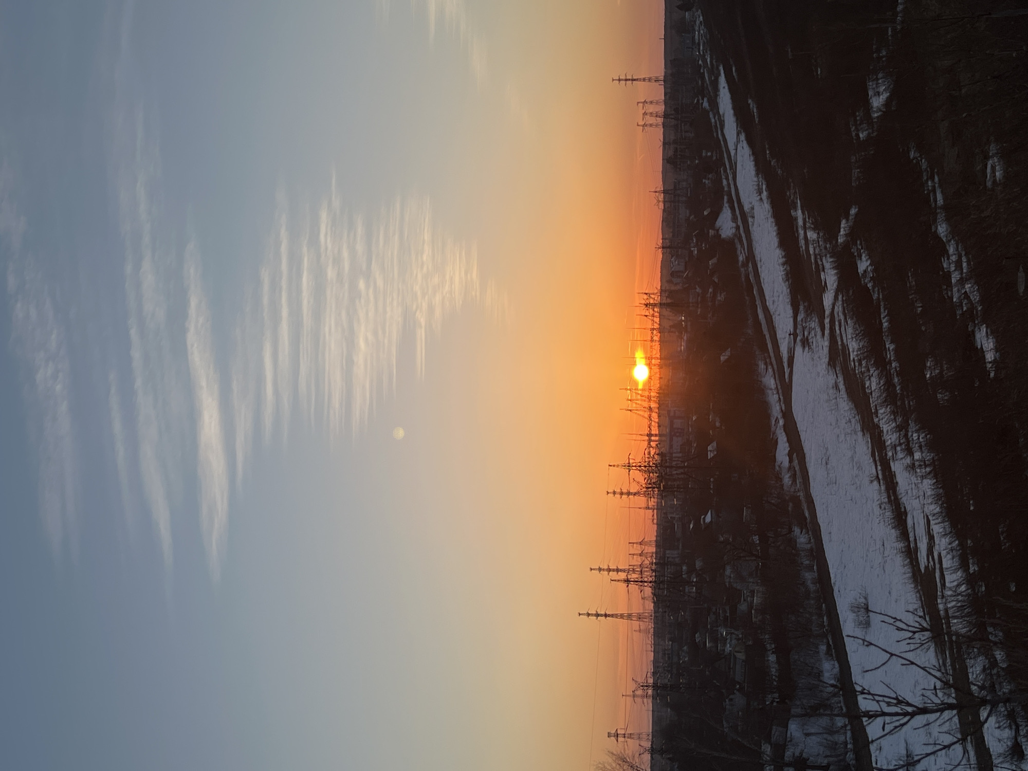 The image captures a sunrise over the industrial city of Chaikovsky, Russia. The sun is low on the horizon, radiating a vibrant orange glow that transitions to a pale blue sky with streaky clouds. In the foreground, silhouetted against the sunrise, are rows of power lines and the outlines of buildings. To the right, patches of snow cover the ground, hinting at a winter or early spring setting. The horizon is somewhat hazy, obscuring distant details. There are no people visible in the shot. The scene is dominated by the industrial infrastructure juxtaposed with the natural beauty of the sunrise.