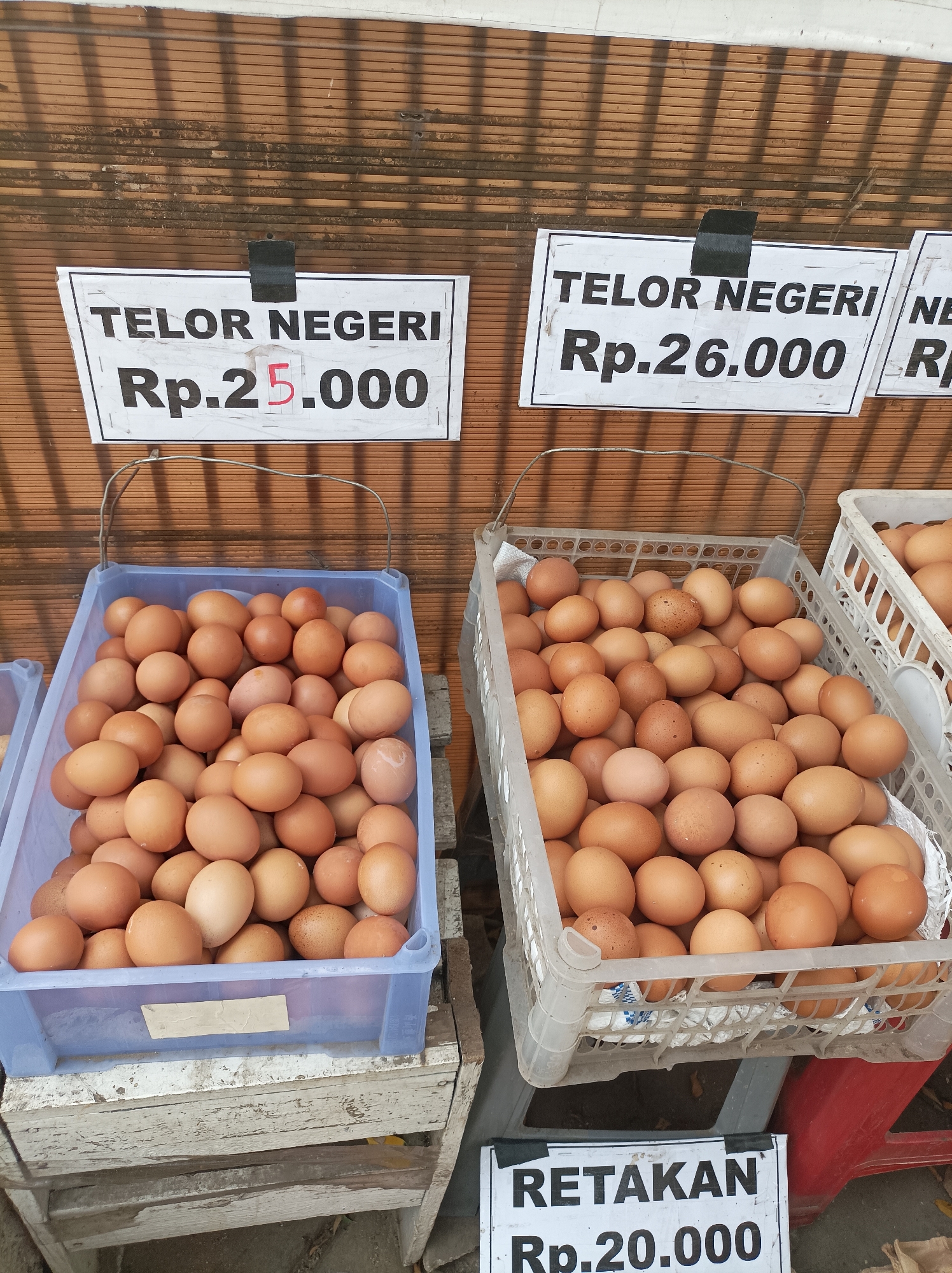 This is a photograph of a street market stall selling eggs in West Jakarta, Indonesia. The stall has several crates of brown eggs, with price signs displayed above them. One sign reads "TELOR NEGERI Rp. 25.000", another "TELOR NEGERI Rp. 26.000", and a third, smaller sign at the bottom reads "RETAKAN Rp.20.000". The background is a textured wall, possibly a building or fence, with shadows cast on it. The lighting suggests it is daytime. There are no people visible in the photograph. The overall scene depicts a commercial transaction point for eggs in an outdoor setting.