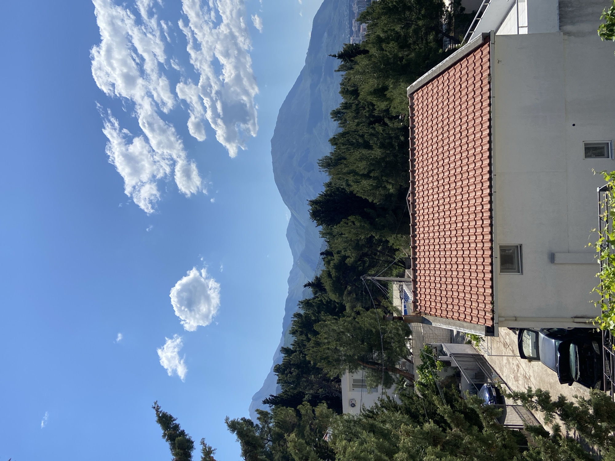 The image captures a view in Šušanj, Montenegro. A bright blue sky with scattered cumulus clouds dominates the left side of the frame. In the background, a large mountain range stretches across the horizon, partially obscured by lush green trees. A building with a terracotta tiled roof occupies the right side of the image, alongside the car parked on the driveway. The angle suggests the shot was taken from an elevated position, possibly a balcony or higher ground, looking out towards the mountainous landscape. The scene appears to be captured on a sunny day, with clear visibility.