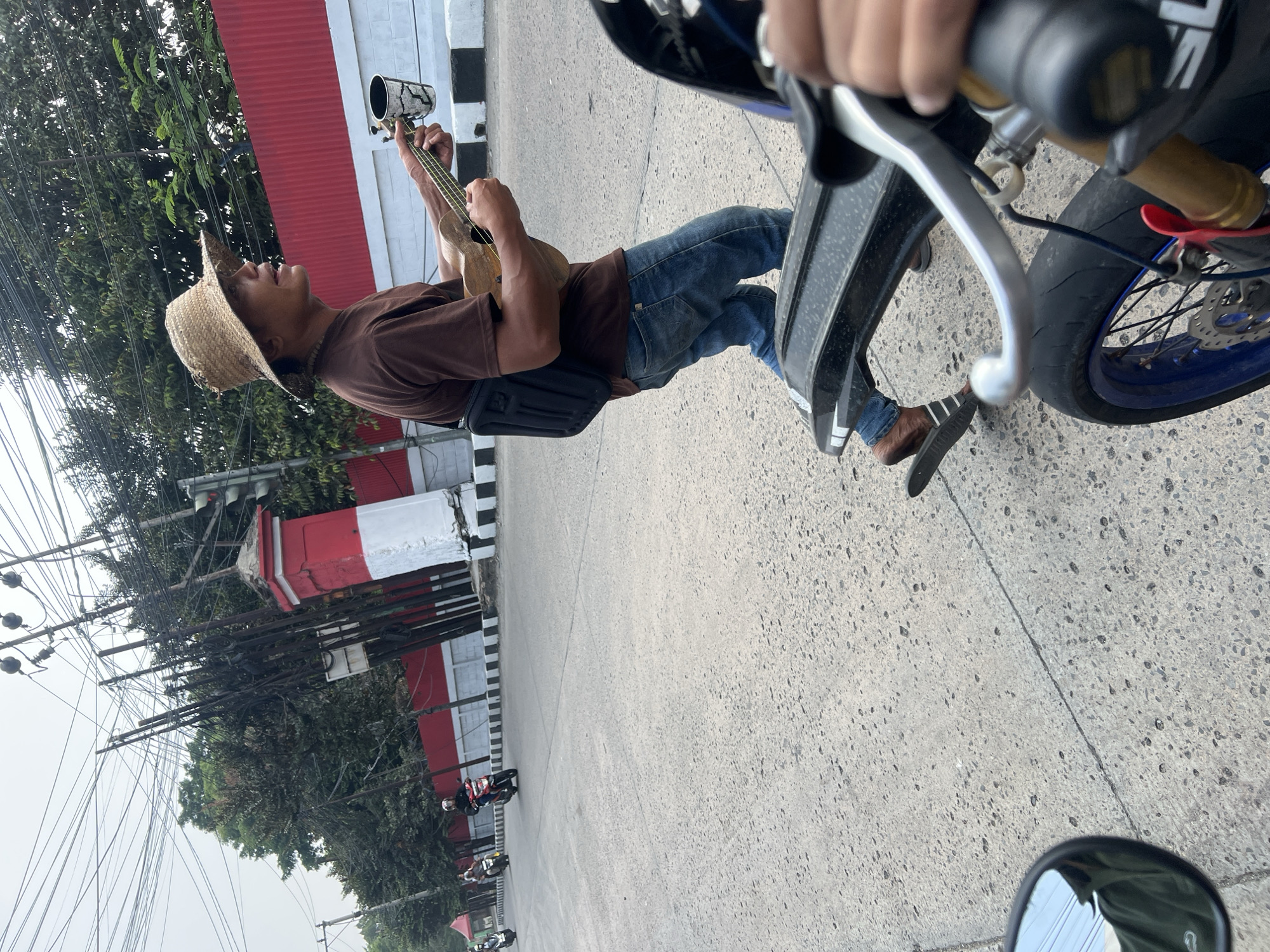 A man wearing a straw hat, brown t-shirt, and blue jeans walks across a paved road, playing a ukulele and holding a cup for donations. He is walking in the direction of a red and white striped pillar and a red wall. Several motorcycles are visible on the road, with some riders wearing helmets. The scene appears to be on a street in Citeureup, Indonesia, with utility poles and wires overhead and trees lining the background. The perspective suggests the photo was taken from a moving vehicle.