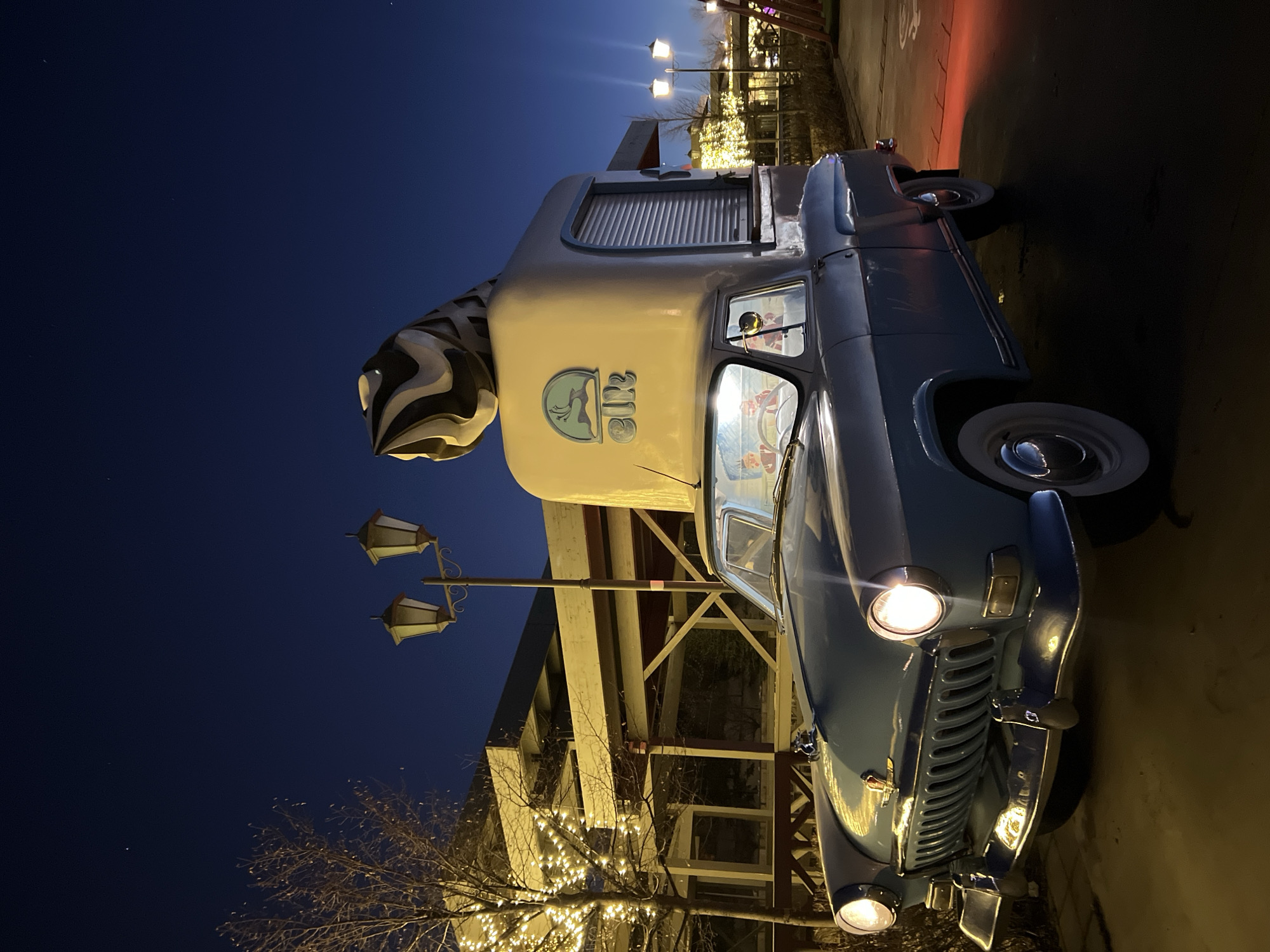 The image depicts a vintage light-blue ice cream truck parked on a paved surface at night, in what appears to be Kazan, Russia. The truck has a large ice cream cone sculpture on top and is illuminated by its headlights and surrounding streetlights. The vehicle is adorned with the logo "Эlli's" and details are visible through the front windshield. In the background, buildings are decorated with strings of lights, suggesting a festive atmosphere. There are vintage style lampposts visible along with foliage that is visible in the darkness.