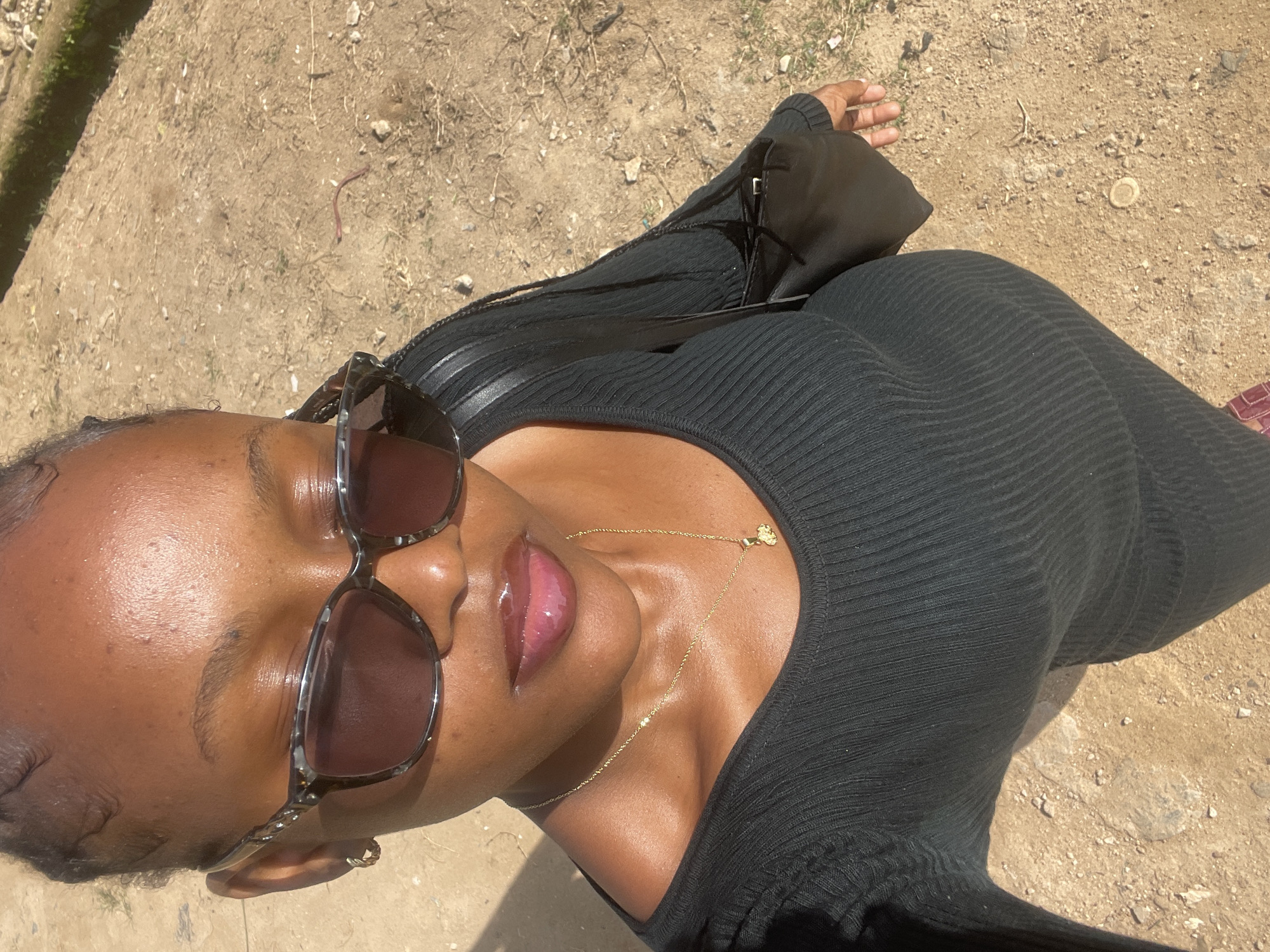 A medium shot captures a young Black woman with braided hair and sunglasses taking a selfie outdoors. She is wearing a black ribbed dress and a gold necklace. Her lips are glossy. The background is a dry, sandy ground with some small stones and dry vegetation. The overall lighting suggests a sunny day. The image is not taken in Jos, Nigeria.