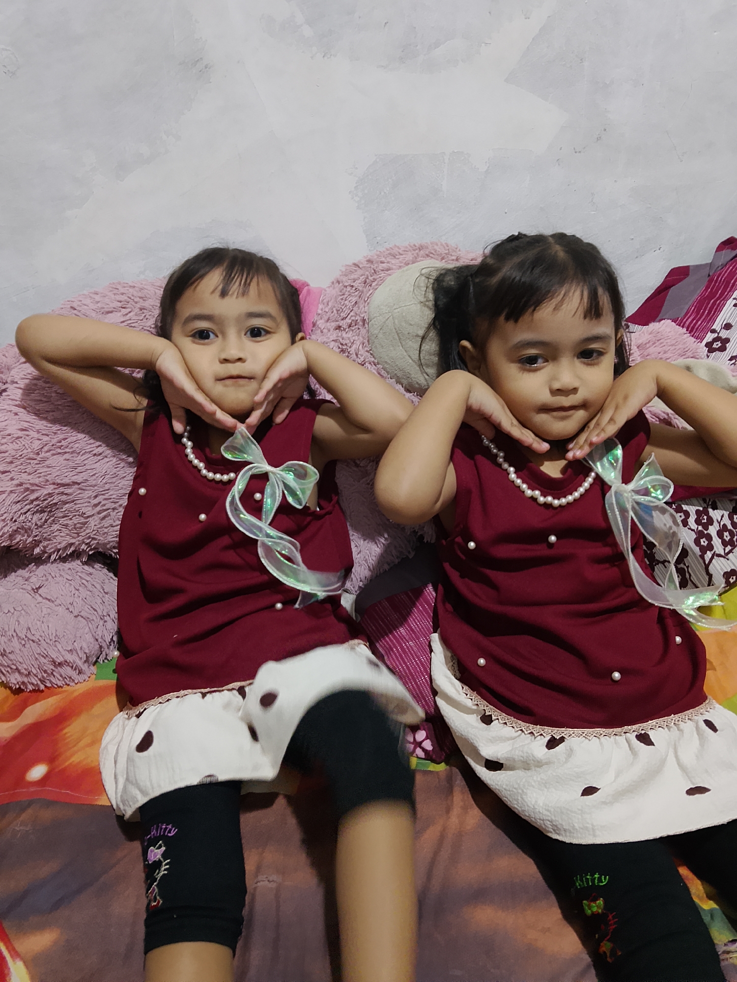 Two young girls, likely sisters, are posing for a photo while lying down on a bed. They are wearing matching maroon sleeveless dresses adorned with faux pearls and a white ribbon bow. The dresses have a white overlay skirt with dark brown polka dots. Both girls have dark hair and are holding their hands to their cheeks in a playful pose. They are wearing black leggings with colorful designs, including what appears to be "Kitty" written on the girl on the right.
The background shows a textured wall and a large, soft pink stuffed animal. The bed is covered with colorful bedding and a patterned blanket. The overall scene suggests a casual, indoor setting. The lighting appears to be from an artificial source, possibly a room light or flash, creating a soft glow. The girls' expressions are alert and engaged, suggesting they are enjoying the photo opportunity.
No specific time of day or weather conditions are discernible from the image. The location context provided indicates Tanggungan, Indonesia.