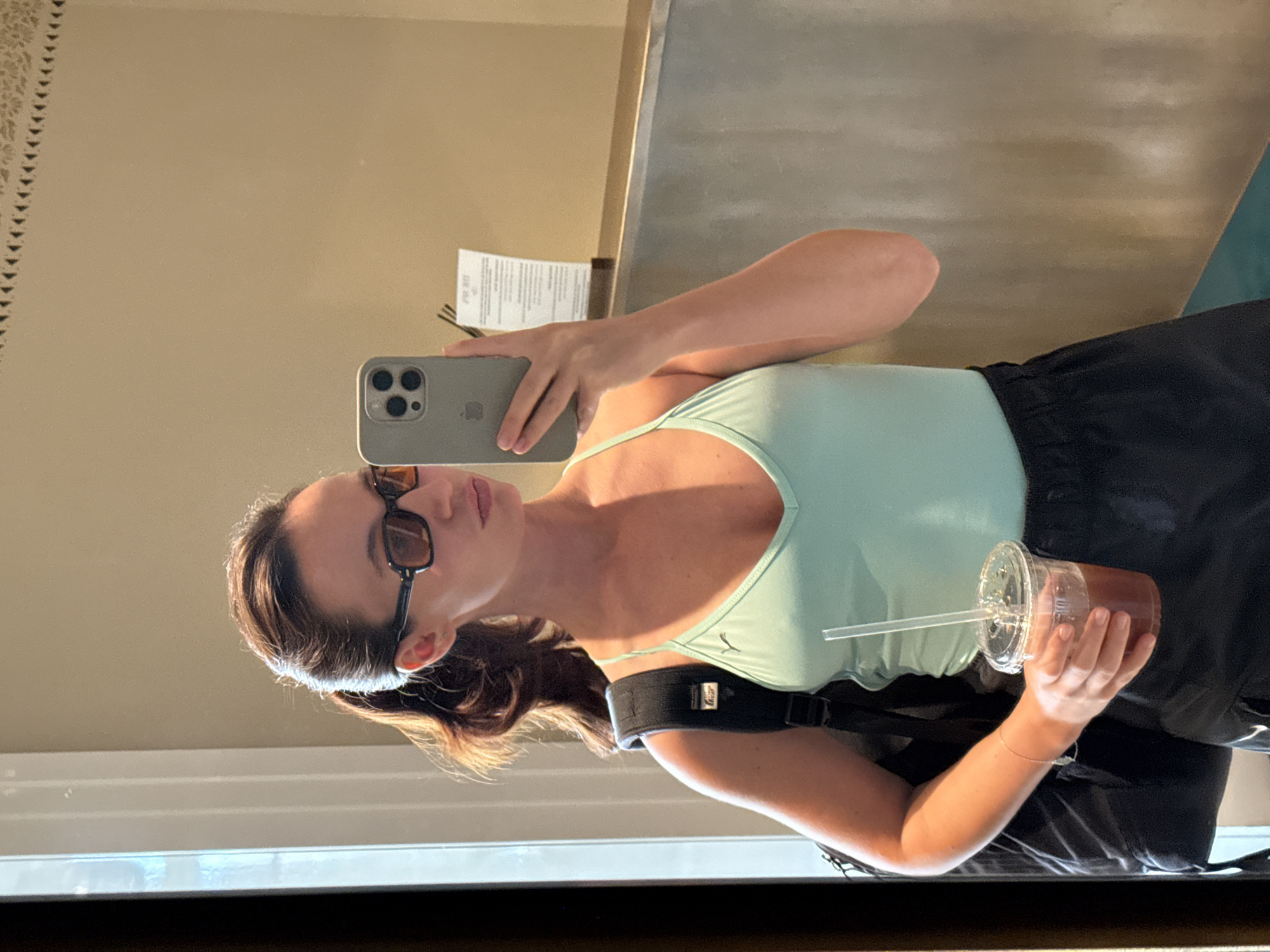 The image shows a woman taking a mirror selfie. She is wearing sunglasses, a light blue tank top, and black shorts. She has a black backpack on and is holding a clear plastic cup with a straw containing a dark beverage. Her hair is dark brown and pulled back. The setting appears to be indoors, possibly in a hotel room or a changing room, given the mirror and neutral wall color. The lighting suggests it is daytime. There is a white piece of paper with text visible on the wall behind her. The woman has a neutral expression.