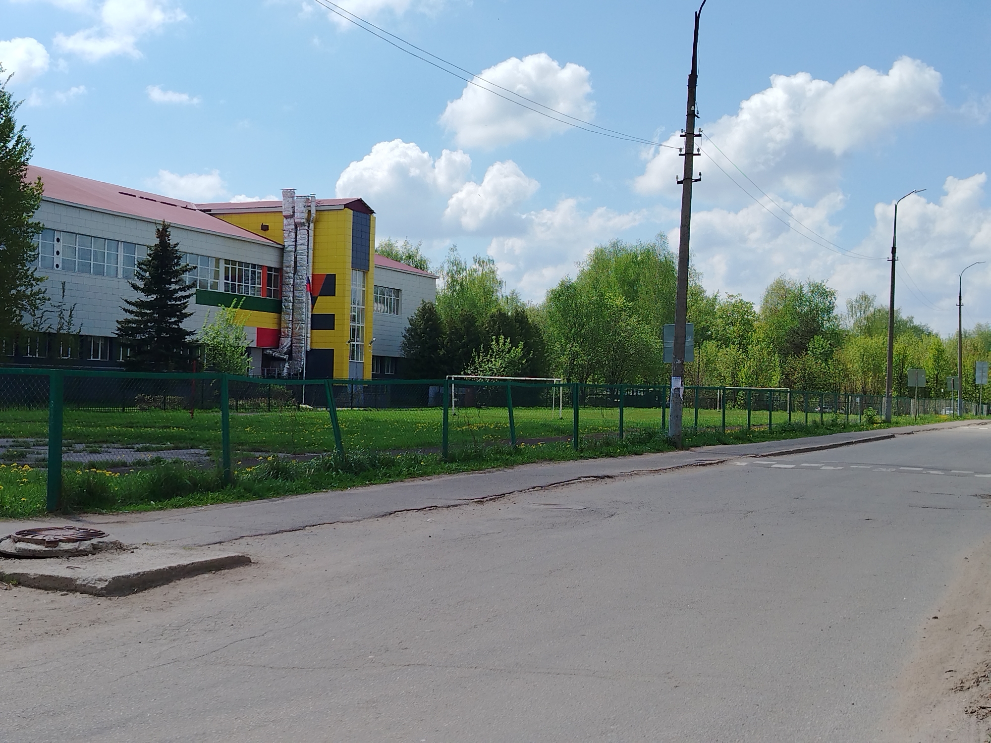 The image depicts a street scene in an urban environment. On the left, there is a multi-story building with a red roof, light-colored walls, and a section with yellow and black geometric designs. A green fence runs along the property line of the building, separating it from the street. Behind the fence is a grassy area with scattered dandelions and a small evergreen tree.
The street is asphalt and appears to be lightly trafficked. On the left side of the street, near the sidewalk, there is a manhole cover surrounded by concrete. The street appears to be at an intersection.
On the right side of the street, there are trees and utility poles. The sky is blue with scattered white clouds. The overall lighting suggests it is daytime. There are no people visible in the scene.