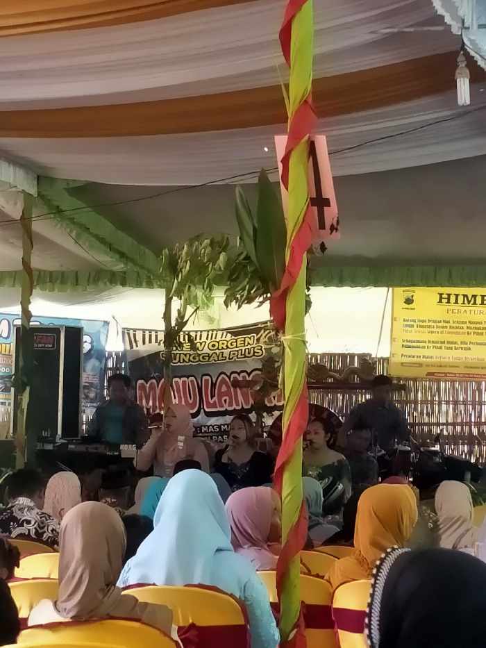 An outdoor event is underway with attendees seated in yellow chairs facing a stage. The stage features a banner that reads "NEW ORGEN TUNGGAL PLUS". Musicians are performing on stage with microphones. Banners and decorations adorn the tent-like structure overhead. A sign on the right contains text and possibly legal information.