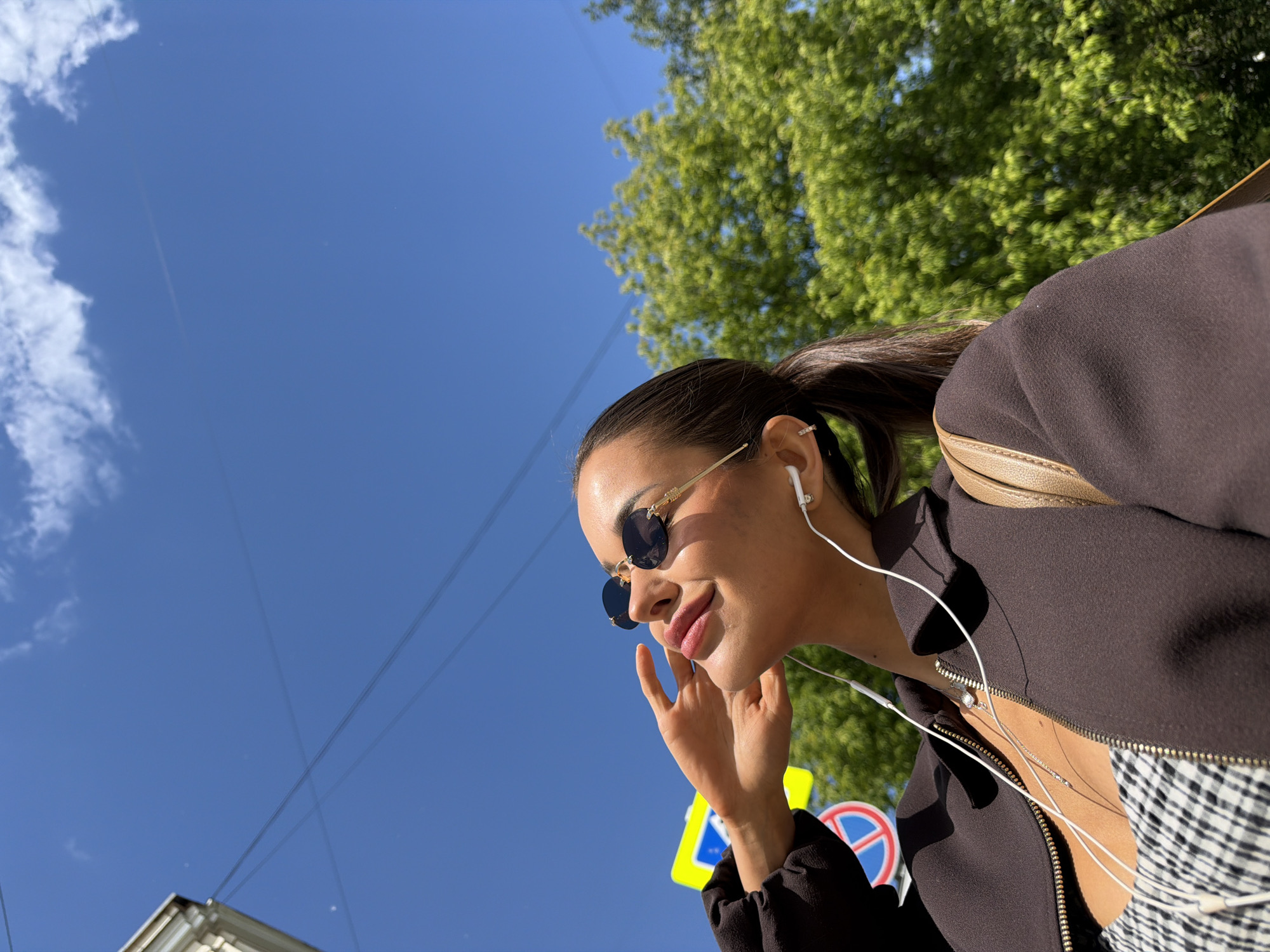This low-angle outdoor shot captures a young woman on a bright, sunny day in Moscow, Russia. She is positioned in the lower right of the frame, looking slightly downwards and to her left with a relaxed, gentle smile, her hand near her chin. Her dark hair is pulled back into a ponytail, and she wears small, round, dark-lensed sunglasses with gold frames. White wired earbuds are in her ears, with the wires extending downwards. She is dressed in a dark brown or black jacket, partially unzipped to reveal a glimpse of a black and white checkered top. A light brown or gold bag strap is visible over her right shoulder.
The setting features a vibrant blue sky, largely clear except for a few wispy white clouds on the left. Lush green tree foliage occupies the upper right portion of the background, suggesting a park or street with trees. Overhead power lines crisscross the sky. In the lower left corner, a partial view of a light-colored building is visible. Below the woman's face, two distinct street signs are present: a yellow square sign and a red circular sign with a white diagonal line, indicative of urban street infrastructure. The image conveys a casual, pleasant moment outdoors under good weather conditions. No visible text is present in the scene.