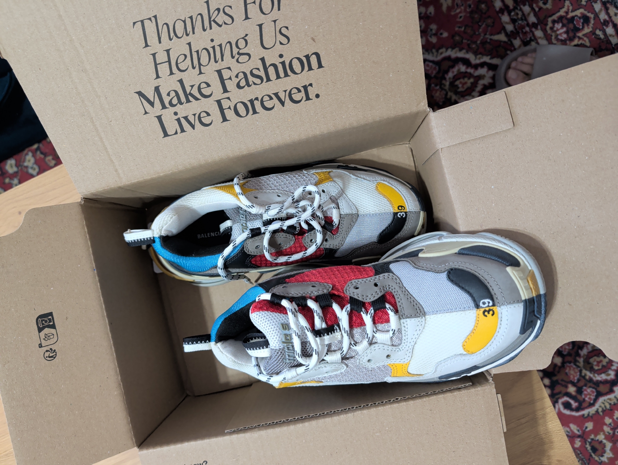The image depicts a pair of multicolored Balenciaga Triple S sneakers contained within an open, light brown corrugated cardboard box, resting on a wooden floor partially covered by a patterned rug. The scene is consistent with an indoor setting in Pleven, Bulgaria.
The left sneaker, oriented towards the viewer, displays a complex design featuring segments of red, blue, yellow, various shades of grey, black, and white mesh and synthetic leather. "Triple S" is visible on its tongue, and the number "39" is printed in black on a yellow segment near the toe. Its laces are white with black accents. The right sneaker, positioned further back and slightly to the right, exhibits a similar multicolor scheme, with "BALENCIAGA" partially visible on its side. Its toe also displays the number "39" on a yellow segment.
The inside flap of the cardboard box, visible on the right, features the printed text: "Thanks For Helping Us Make Fashion Live Forever." The top-left inner flap displays recycling symbols, including a figure dropping waste, a recycling bin, and a coffee cup recycling icon. The floor beneath the box consists of light-toned wooden planks and a segment of a dark red rug with intricate beige and black patterns. A human foot, wearing a light-colored sandal, is partially visible in the bottom right corner of the frame, positioned on the rug next to the box.