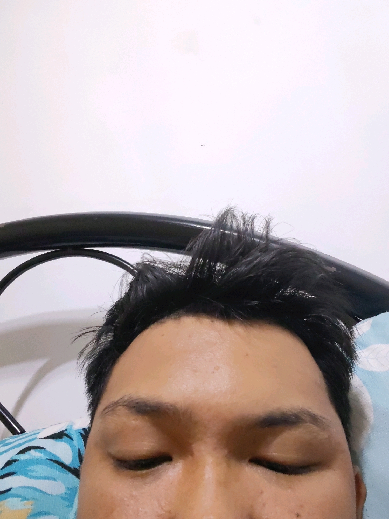 A close-up, eye-level, medium shot captures the top portion of a person's face, specifically their forehead, eyebrows, and closed eyes. The individual has dark, messy hair that is slightly disheveled. They are lying down, as indicated by the presence of a pillow with a blue and white patterned fabric beneath their head and a black metal bed frame arching above their hair. The background is a plain white wall. The lighting appears to be from overhead. The scene is indoors, suggesting a bedroom setting, likely in Naic, Philippines, given the context.