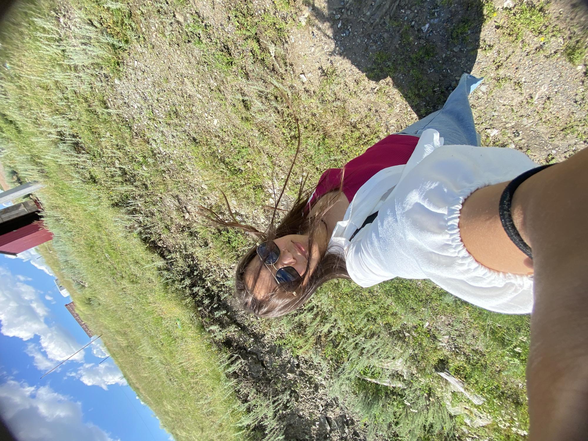 In Yuldybayevo, Russia, a young woman is captured in a selfie. She is wearing sunglasses, a white shirt, and a pink top. Her hair is dark and windswept. The background is a grassy hillside with sparse vegetation and rocky ground. A red building can be seen in the distance on the top of the hill. The sky is blue with scattered white clouds. The image is taken outdoors on a sunny day.