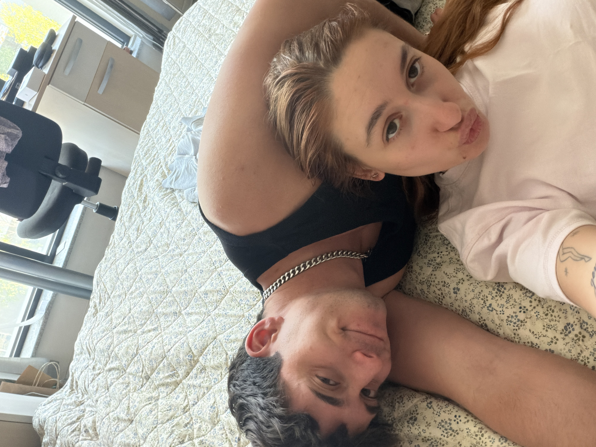 An overhead shot captures a man and a woman lying close together on a bed, possibly taking a selfie. The woman, with reddish-brown hair, is positioned on her back, looking directly at the camera with her lips playfully pursed. She is wearing a light-colored, possibly pink, t-shirt, and a tattoo of a figure and a star is visible on her left arm. The man, with dark, curly hair and a silver-colored chain necklace, lies beside her, resting his head on her arm or chest, and looks towards the camera with a subtle, gentle smile. He is wearing a black top.
They are resting on a light-colored, quilted bedspread adorned with a subtle floral or patterned design in hues of green and blue. The setting appears to be an indoor room, likely a bedroom or living space, in Sofia, Bulgaria, as indicated by the provided context. Natural light streams in from a window visible in the background, suggesting it is daytime. An office chair with a black backrest and a light-colored cabinet or set of drawers are partially visible further back in the room. A couple of brown paper bags are placed on a window sill or surface near the window. The overall mood conveyed by their expressions is relaxed, affectionate, and contented. There is no visible text in the image.