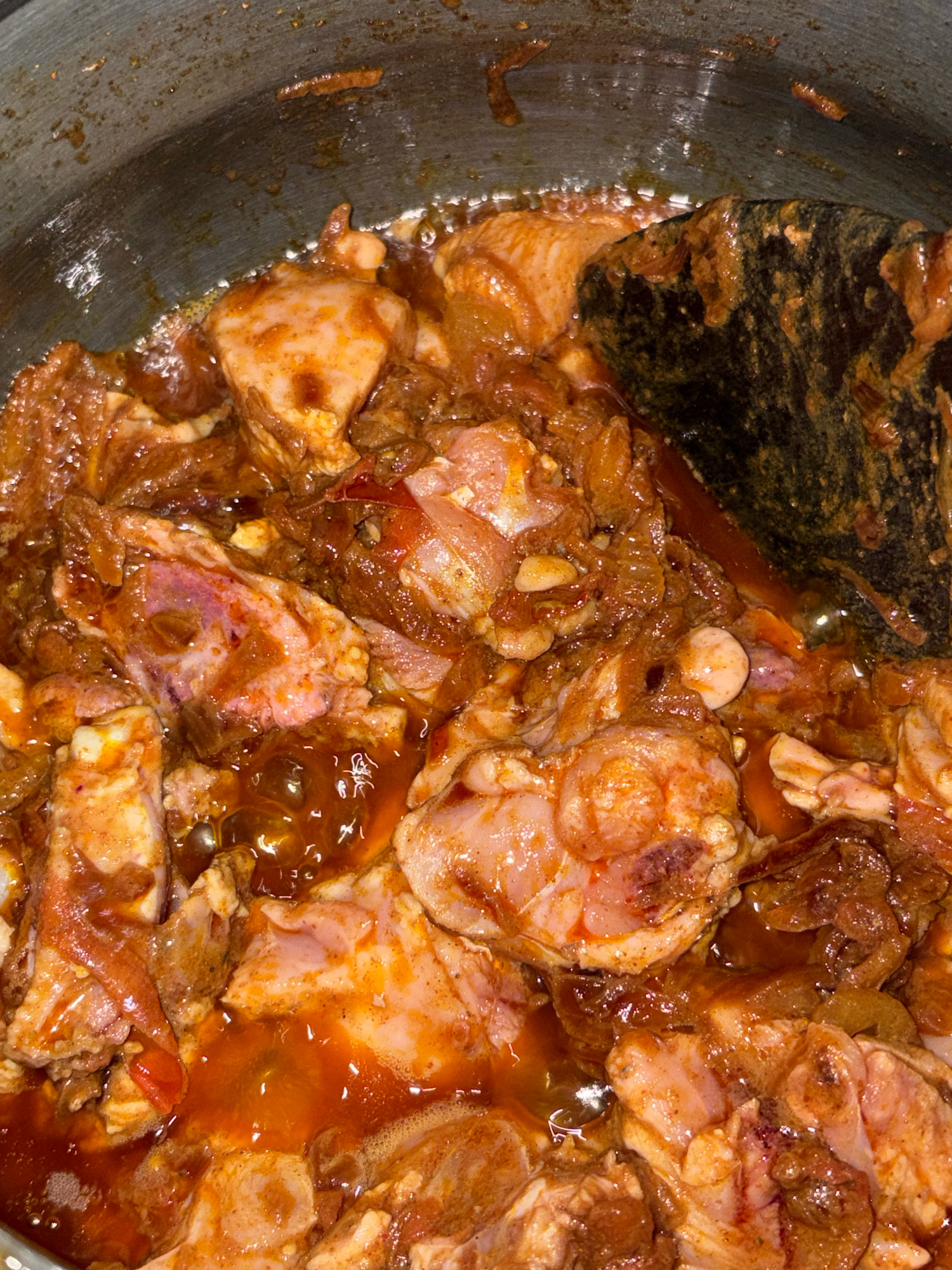A close-up, overhead shot shows pieces of raw chicken being cooked in a dark-colored pot with a wooden spoon resting inside. The chicken is coated in a reddish-orange sauce that appears to be simmering. There are some chopped onions and other ingredients mixed in with the chicken. The pot's interior is slightly greasy, and some splatters of the sauce are visible on the rim. The lighting is dim, creating a warm and rustic feel.