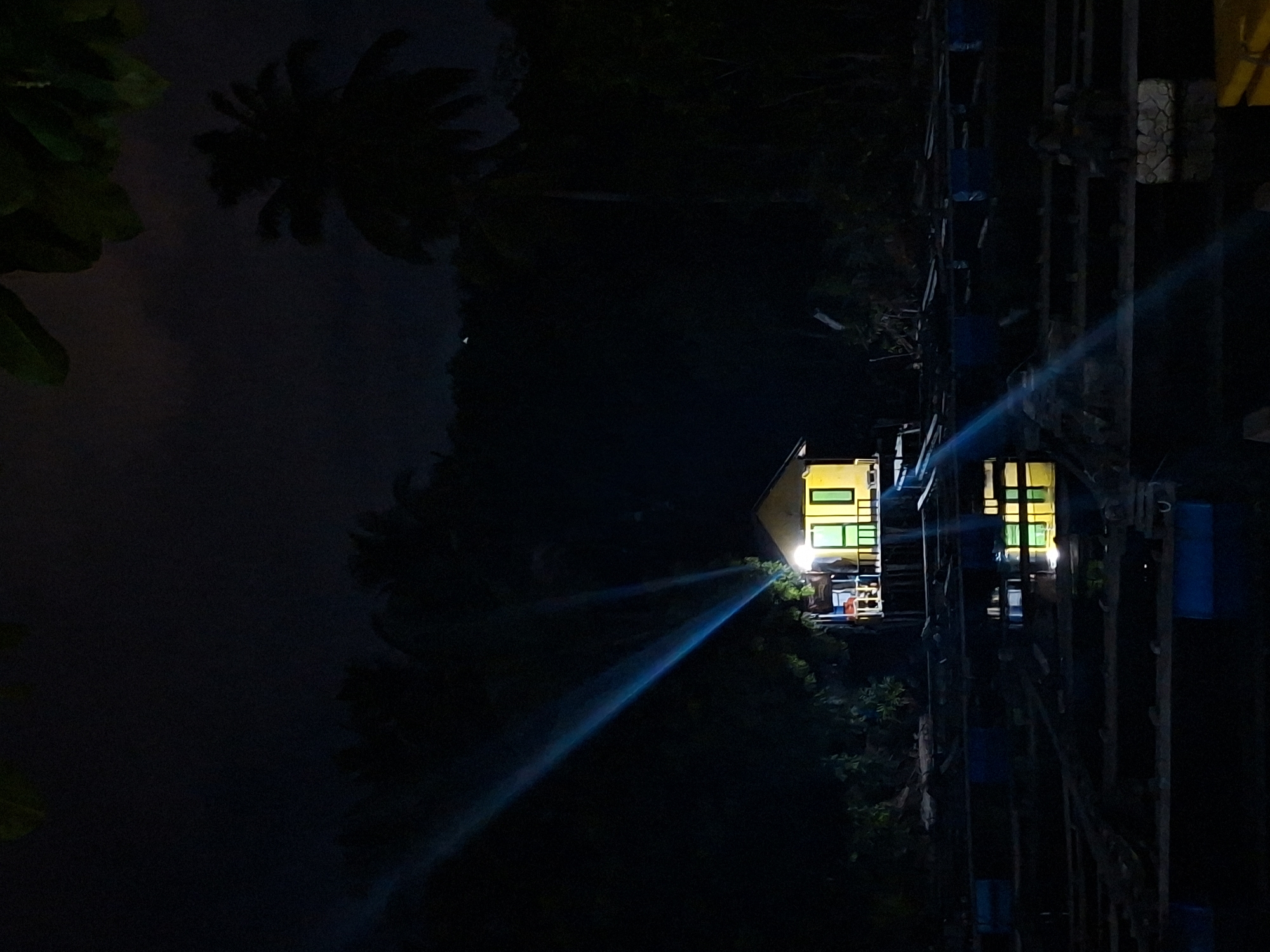 The image shows a dimly lit scene at night in Lhokseumawe, Indonesia. A two-story yellow building with bright interior lights stands amidst dark vegetation. A bright blue beam of light projects from the building toward a dark, potentially watery area. Adjacent to the building is a structure of metal bars and blue containers. The overall environment appears to be a mix of urban and natural elements at night.