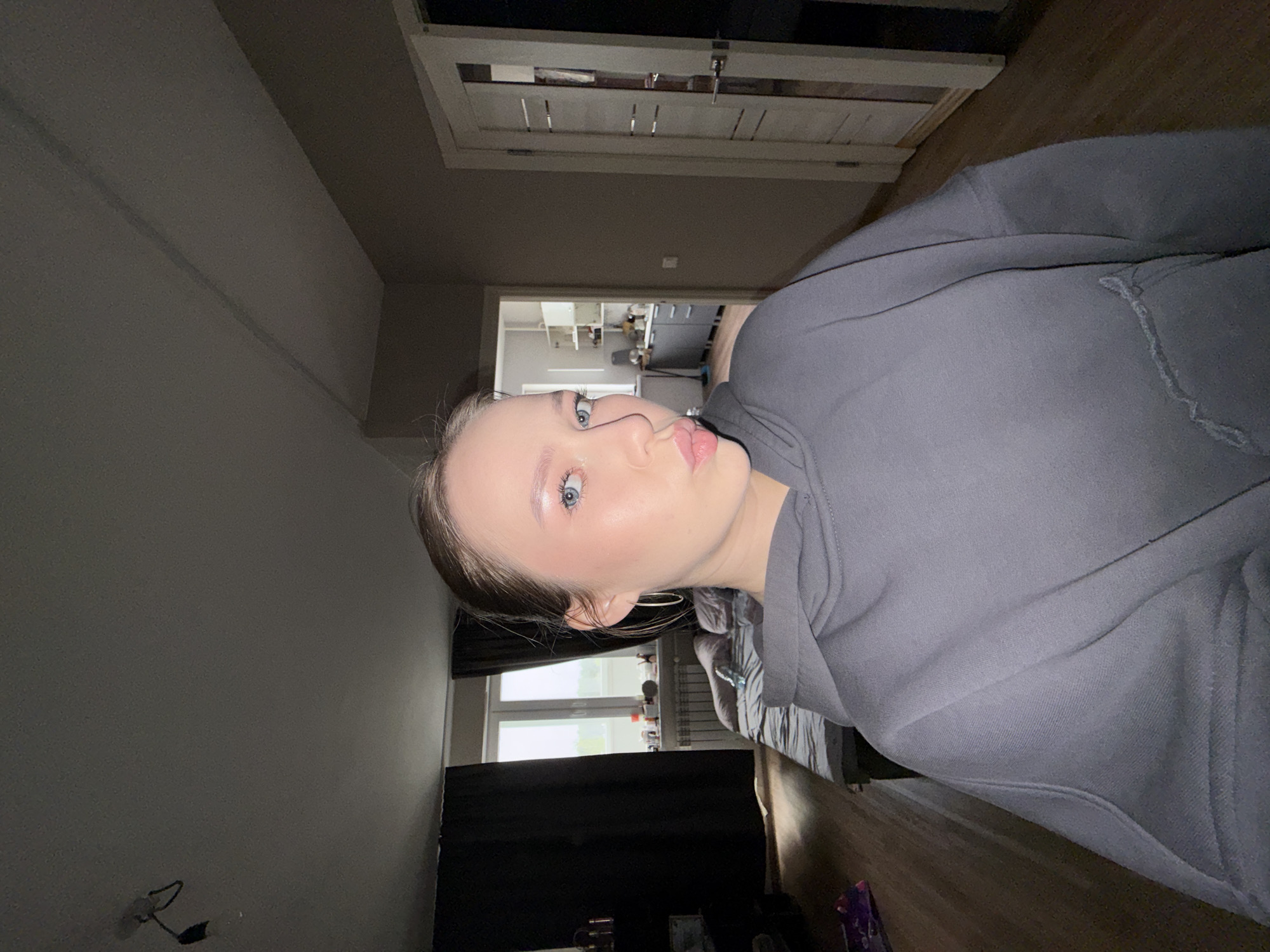 A young woman is captured in a medium close-up, posing indoors in what appears to be a home setting. She has fair skin, prominent blue eyes, and light brown hair, which is styled back. Her face features light makeup, including subtle eyeshadow, blush, and lip gloss, enhancing her features. She is wearing a comfortable grey hooded sweatshirt and a simple hoop earring on her visible ear. Her expression is composed, with slightly parted lips, directed towards the camera.
The background reveals a multi-room domestic interior. To the woman's left, a white door with a silver handle and lock mechanism is clearly visible. Beyond this, a doorway opens into what looks like another room, possibly a kitchen or utility area, with white cabinets, shelving, and indistinct appliances visible. To her right, a window with a white frame provides some natural light, although it is partly covered by dark curtains. A section of a bed with grey bedding can be seen in the lower right portion of the image. The flooring throughout the visible areas is made of wood. On the wall to the far left, an electrical outlet with some exposed wires is present. The overall lighting is subdued, suggesting either indoor artificial lighting or a dimly lit time of day. No text is visible in the image. The scene depicts a casual moment within a residence located in Chaikovsky, Russia.