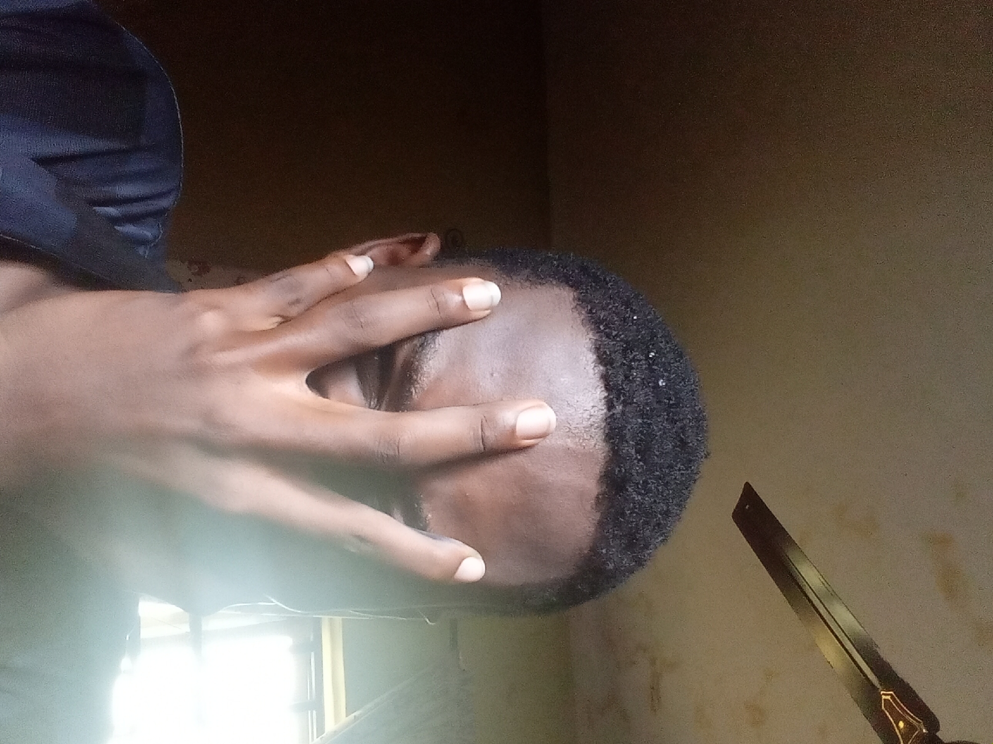 The image displays a close-up, rotated portrait of a person, likely male, with dark skin and short, dark, tightly curled hair. His right hand is prominently positioned, covering the upper half of his face, including his eyes and forehead, with his fingers spread across his brow. His fingernails are light-colored. He appears to be wearing a dark blue or black garment, visible on the upper-left side of the frame.
The setting is indoors, featuring plain, light brown or tan walls that exhibit some minor discoloration or texture. A strong, bright light source, likely a window, is visible in the lower-left corner, creating a significant glare. This indicates the photo was taken during the day.
In the bottom-right corner, a distinctive object is partially visible. It is a bladed tool, possibly a knife or a short machete, with a dark metallic blade and a handle adorned with ornate brown and golden accents.
The person's act of covering their face could imply various interpretations, such as shielding their eyes from the bright light, a gesture of shyness, or a moment of reflection. There is no visible text in the image. The provided context of Onitsha, Nigeria, aligns with the subject's appearance.