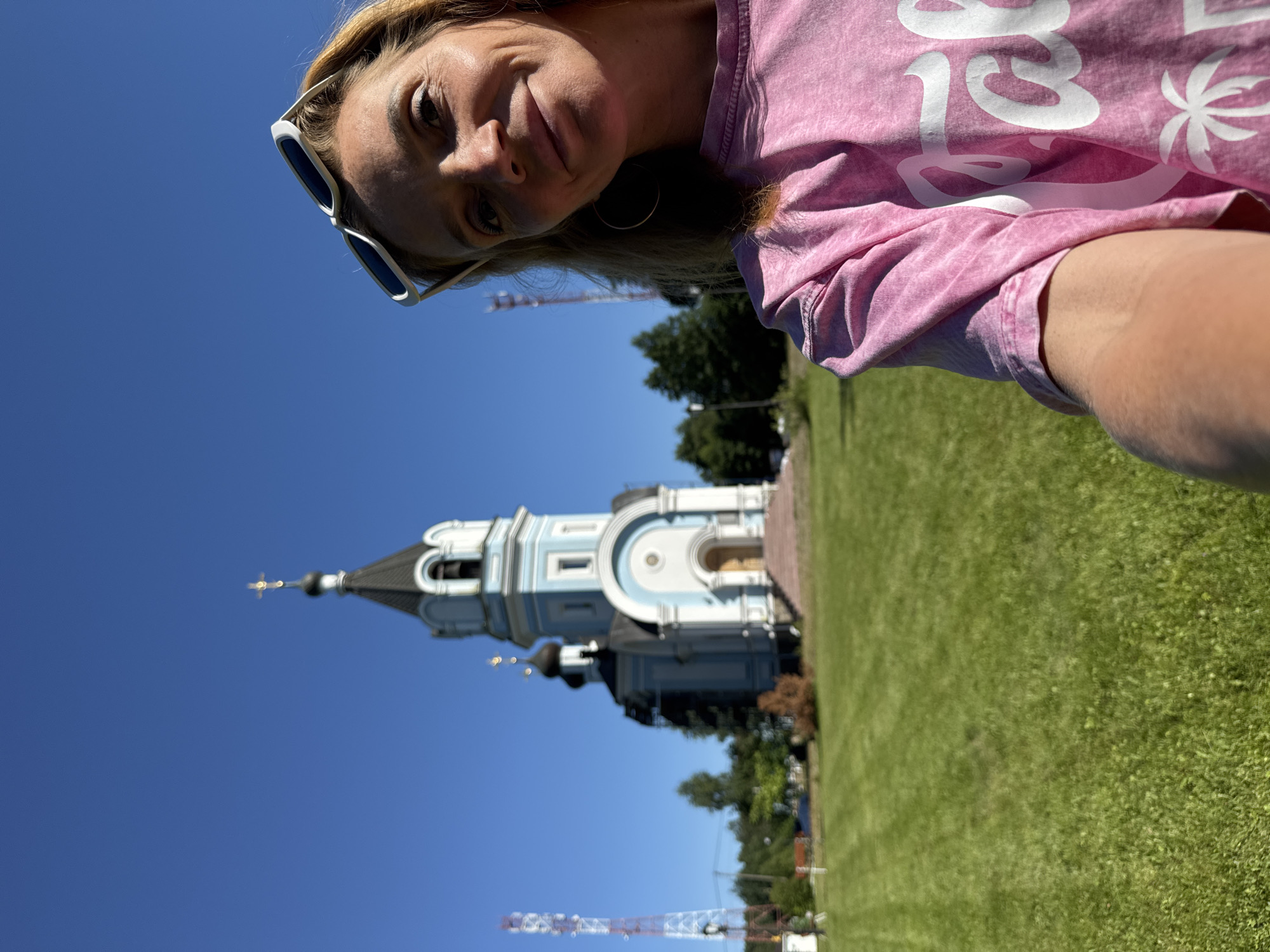 A selfie is taken by a blonde woman with sunglasses perched on her head, smiling at the camera. She wears a pink t-shirt. In the background, a light blue and white orthodox church with a tall steeple stands under a clear blue sky. The church has a black dome with a golden cross on top. In front of the church, a manicured green lawn stretches out. To the left of the church, a tall cell tower with red and white accents is visible. Trees are scattered throughout the scene. The woman appears to be in St. Ozerki, Russia.