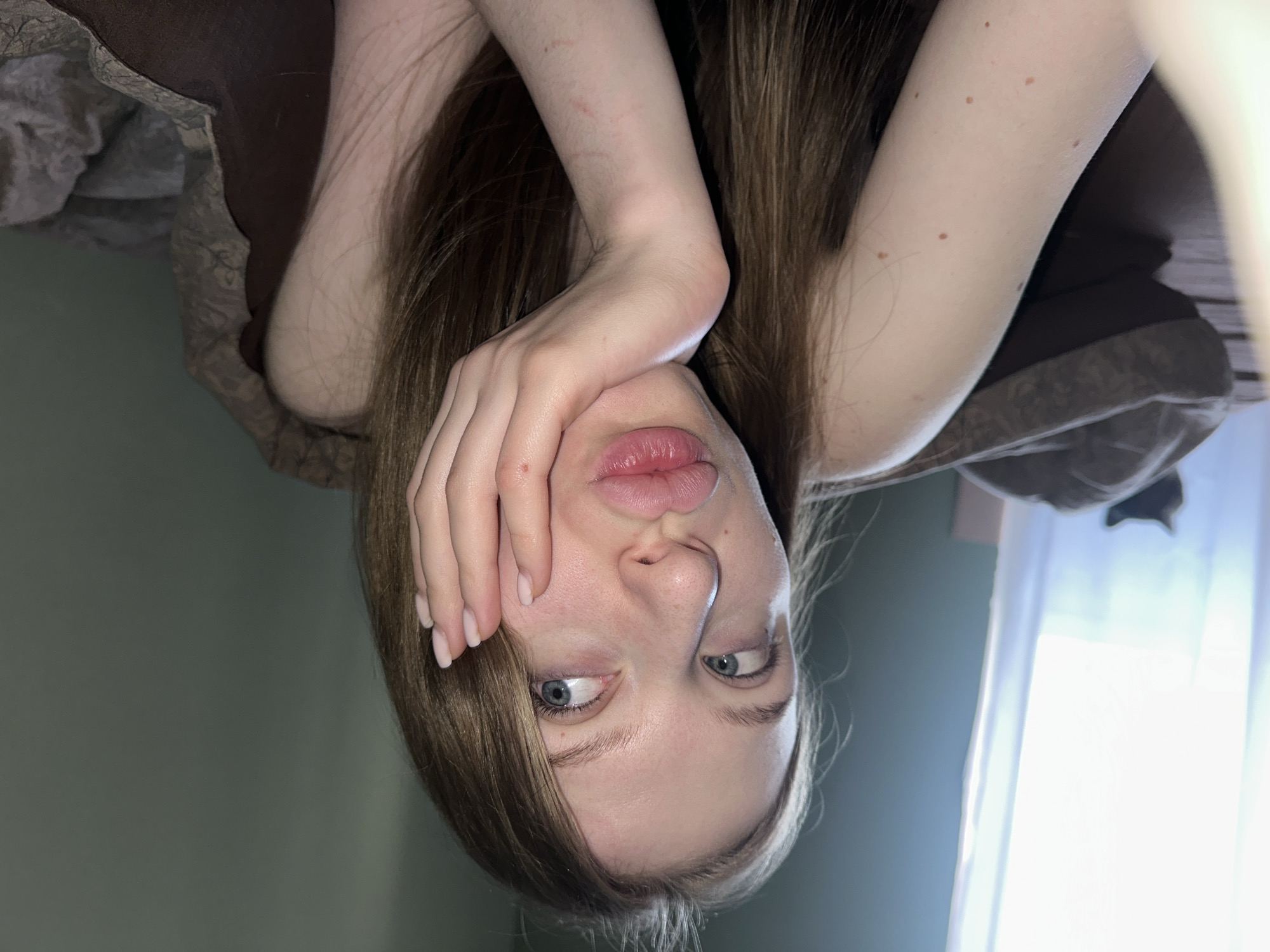 A light-skinned young woman with long, light brown hair is upside down in a room with green walls and brown bedding. She is holding her hand up to her face, covering part of her cheek. Her lips are pursed, and her eyes are slightly crossed. A window with white curtains is visible in the background. The location of the scene is unknown and therefore can not be the city of Chaikovsky, Russia.