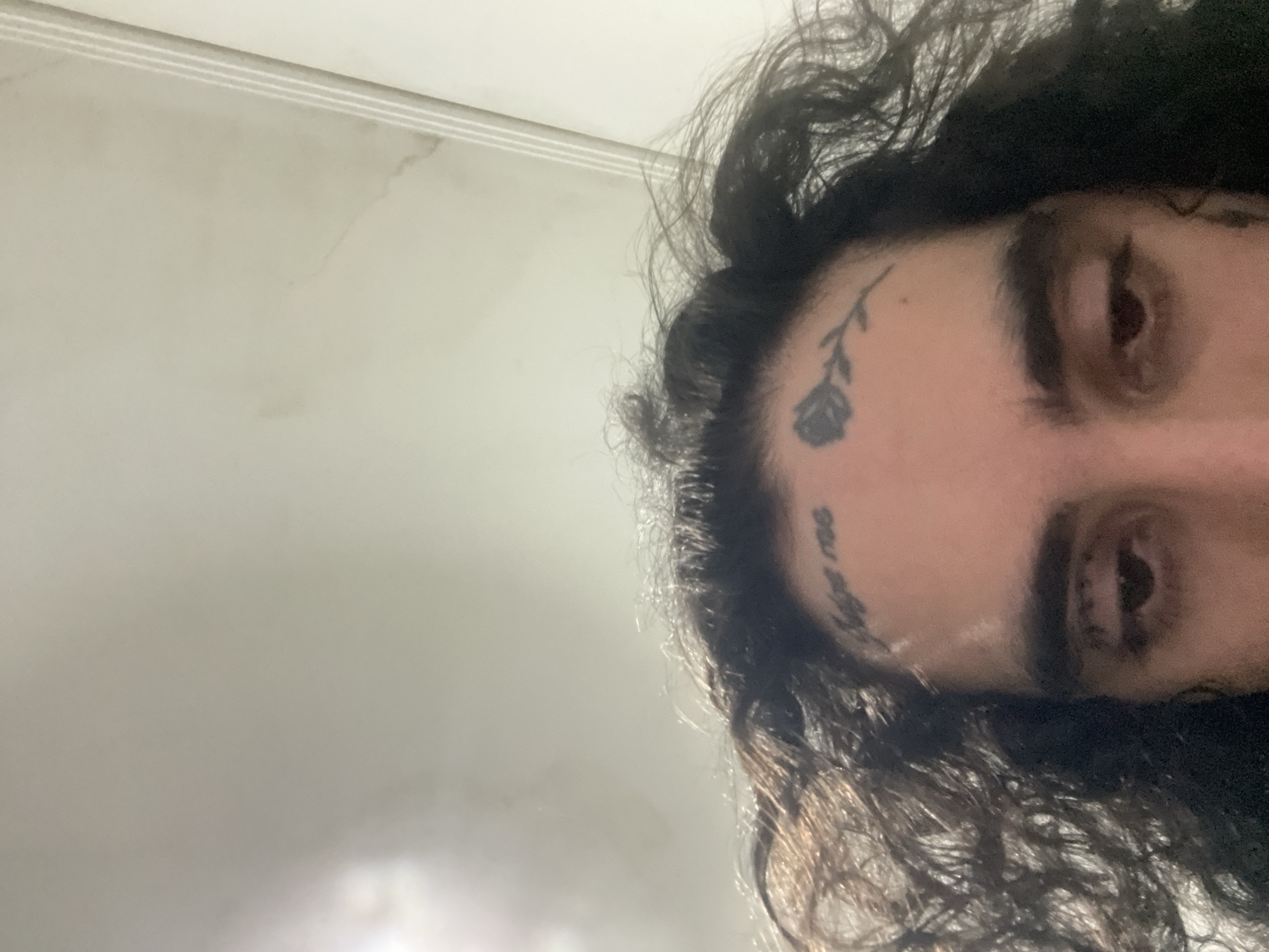 A close-up, angled shot of a person's face. The person has dark, curly hair, visible eyes, and eyebrows. The person has at least two tattoos on their forehead, one a rose and one a word or phrase. The background appears to be a light-colored wall or ceiling.
This image doesn't show enough information to determine a location in Buenos Aires or any other specific place.