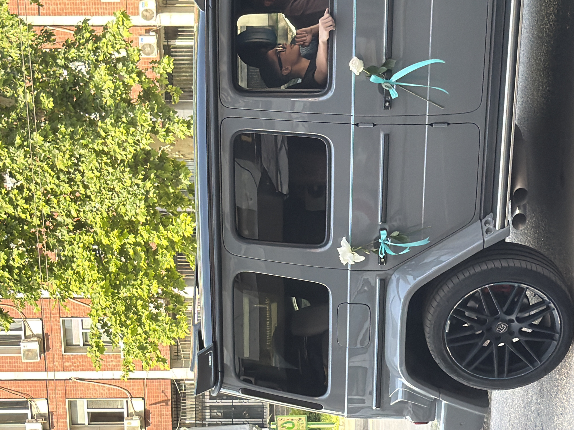 The image shows a gray Mercedes-Benz G-Class SUV, decorated with two white roses and light blue ribbons on its doors. A person with dark hair and sunglasses is leaning out of the front passenger window, looking downwards. The car is parked on a street, possibly in Pleven, Bulgaria, as indicated by visual clues (though not explicitly stated). A tree with dense green leaves is visible to the left, partially obscuring a brick building with air conditioning units on its exterior. The vehicle has aftermarket black rims and visible exhaust pipes. The focus is on the decorated vehicle and the individual in the front seat.