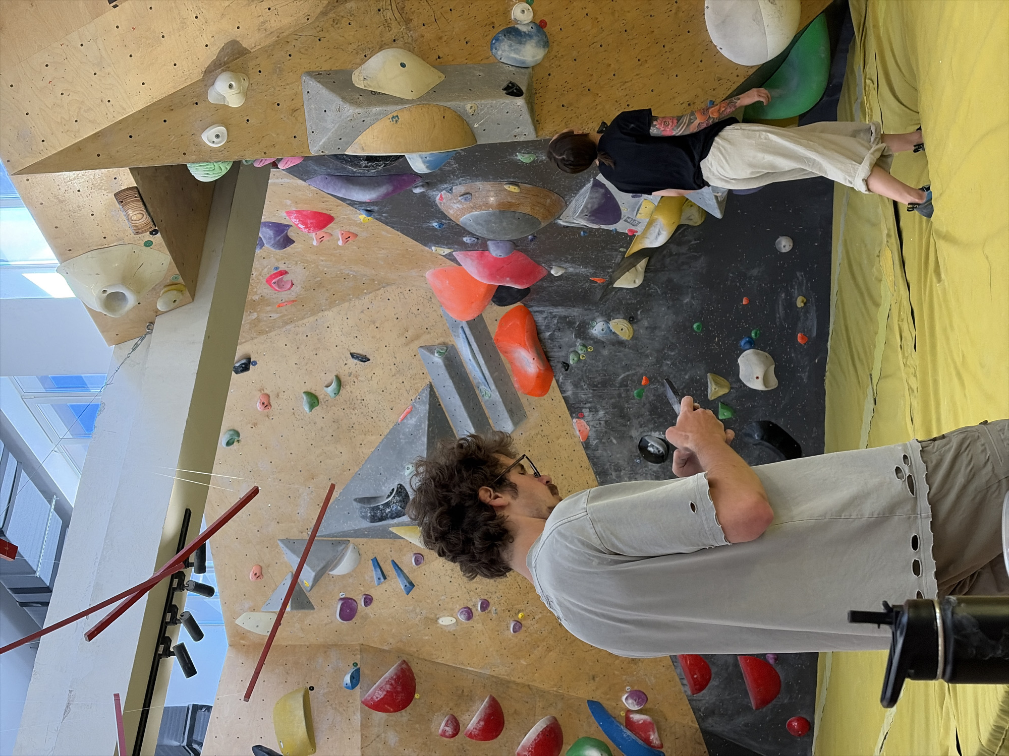 The image depicts an indoor bouldering gym. A man with curly brown hair and glasses is standing in the foreground, facing away from the camera and looking at his phone. He is wearing a light gray t-shirt with ripped details on the sleeves and hem, and khaki shorts. To the right, a person with dark hair pulled back in a ponytail, wearing a black t-shirt and light-colored wide-leg pants, is walking on the crash pad. They have a tattooed arm. The background consists of a large, multi-colored climbing wall with various handholds and structures. The floor is covered with a yellow crash pad. The environment appears to be a modern indoor recreational facility. The specific location within Moscow, Russia cannot be determined from the image.