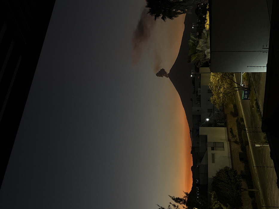 At dusk, a volcano stands tall against an orange-tinged sky. Smoke billows from its peak. Adjacent to the volcano is a row of buildings, their lights puncturing the dimness. Palm trees frame the scene, adding a tropical touch. A road runs alongside the buildings, and a dark foreground adds depth to the composition.