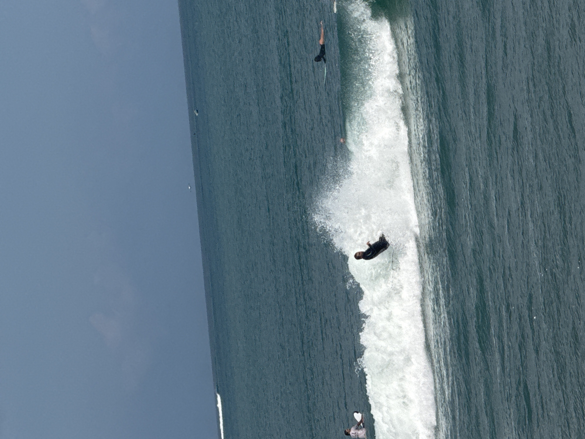 In the ocean off Kuta, Indonesia, surfers ride waves under a clear sky. A surfer in a black wetsuit is actively riding a breaking wave, spray erupting around them. To the left, another surfer in a white shirt and pink shorts is paddling on a surfboard. Further out to sea, another surfer in a black wetsuit lies prone on their board. The water is a deep blue-green, with whitecaps indicating the presence of waves. In the distance, the horizon is visible, with a faint cloud formation in the sky.