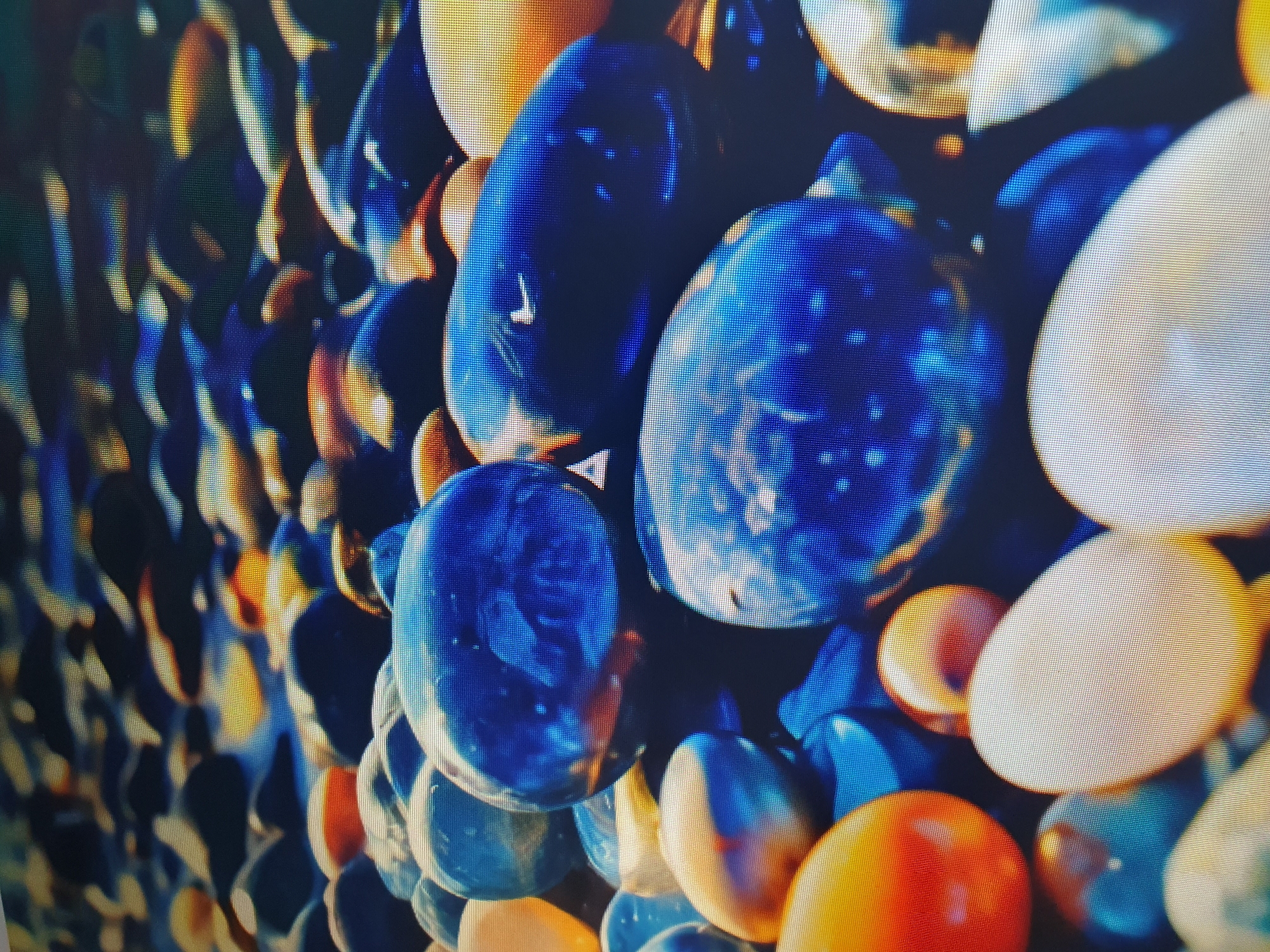 The image is a close-up, low-angle shot of a collection of smooth, colorful pebbles, likely on a beach or a decorative garden feature. The pebbles are varied in size, shape, and color, ranging from deep blues and vibrant oranges to creamy whites and darker tones. The lighting creates reflections on the wet surfaces of the pebbles, giving them a glossy appearance. In the background, the pebbles continue, creating a textured, out-of-focus field. There is no discernible urban environment or specific location within Tashkent, Uzbekistan visible in the image. The focus is entirely on the natural texture and colors of the pebbles.