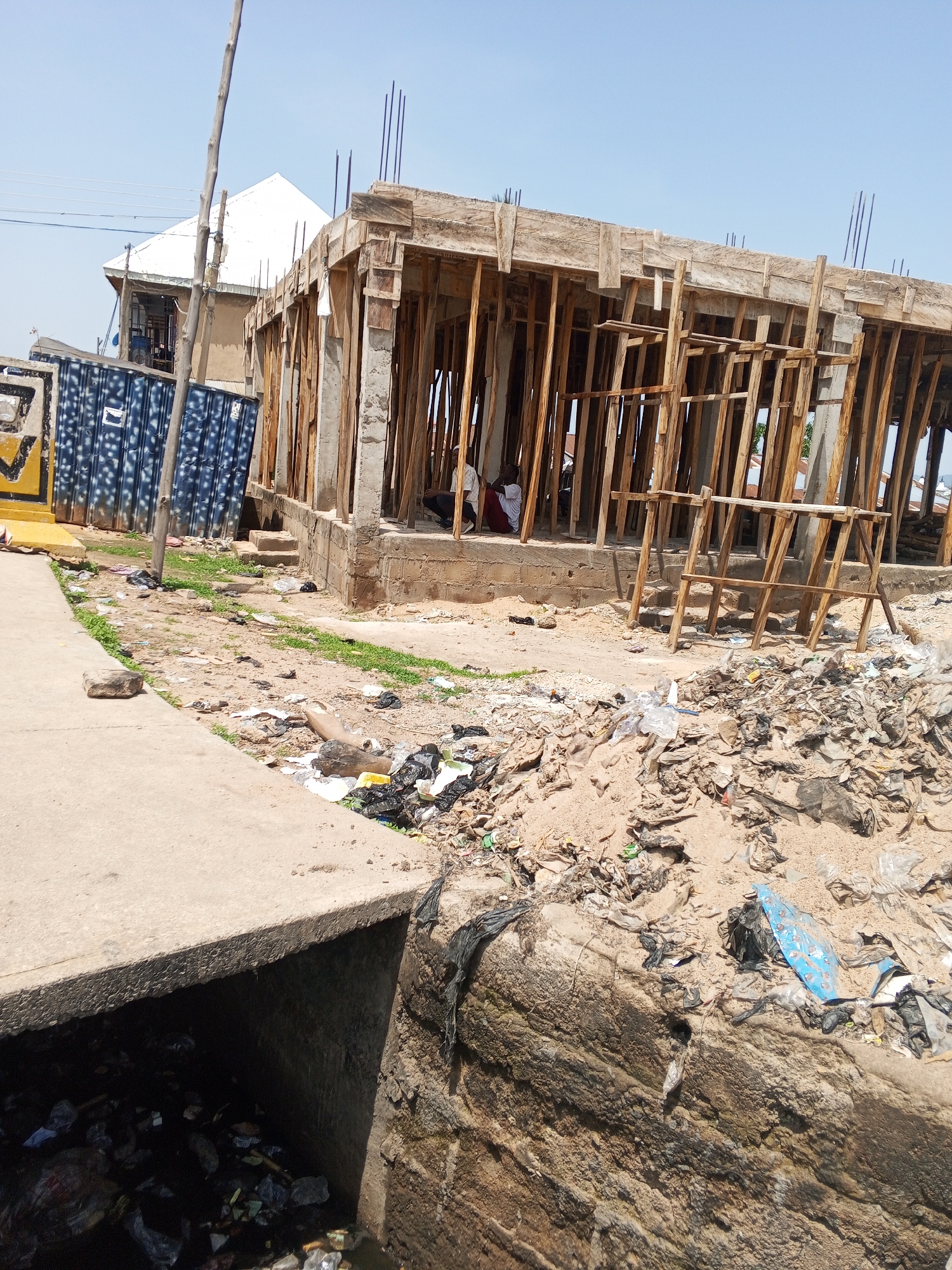 In Jalingo, Nigeria, a construction site is visible, with concrete pillars and wooden framing for a building. In the foreground, a concrete drainage channel is filled with dark water and refuse. Piles of garbage, including plastic bags and debris, are scattered on the ground next to the construction area. A blue metal structure, possibly a shipping container, is situated to the left of the construction site. A person in a white shirt is partially visible inside the building, and another person in dark clothing is seated nearby. The sky is clear and blue.